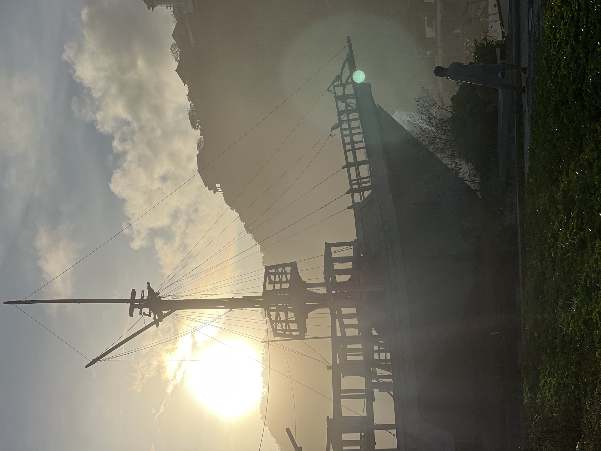 The image captures a maritime scene against a backdrop of a hazy, sunlit sky with some visible clouds. A tall ship, its rigging prominent against the sun, stands as the central subject. The ship's mast extends upwards, while its hull is partly obscured by the bright sunlight. A figure is partially visible walking along a path or walkway near the ship. Lush green vegetation lines the edge of this pathway. The scene is likely located in or near Kuta Selatan, Indonesia, given the prompt information.