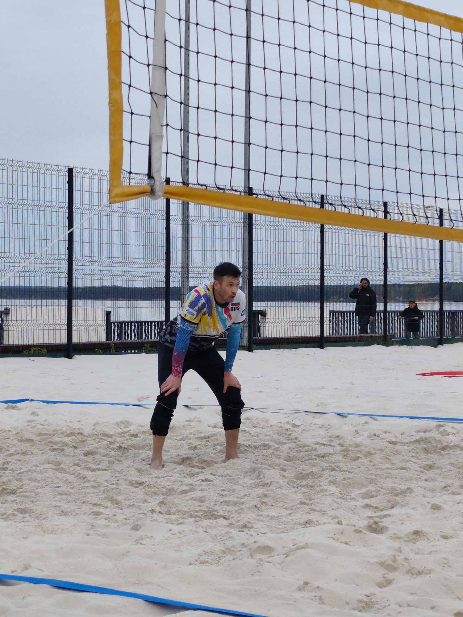 The image shows a man in athletic attire with "RUS" on the sleeve, positioned on a sand volleyball court with his hands on his knees. A volleyball net with yellow trim is prominently displayed behind him. In the background, a fence separates the court from a body of water, with two people visible on a path or pier. The image was taken in the city of Chaikovsky, Russia.