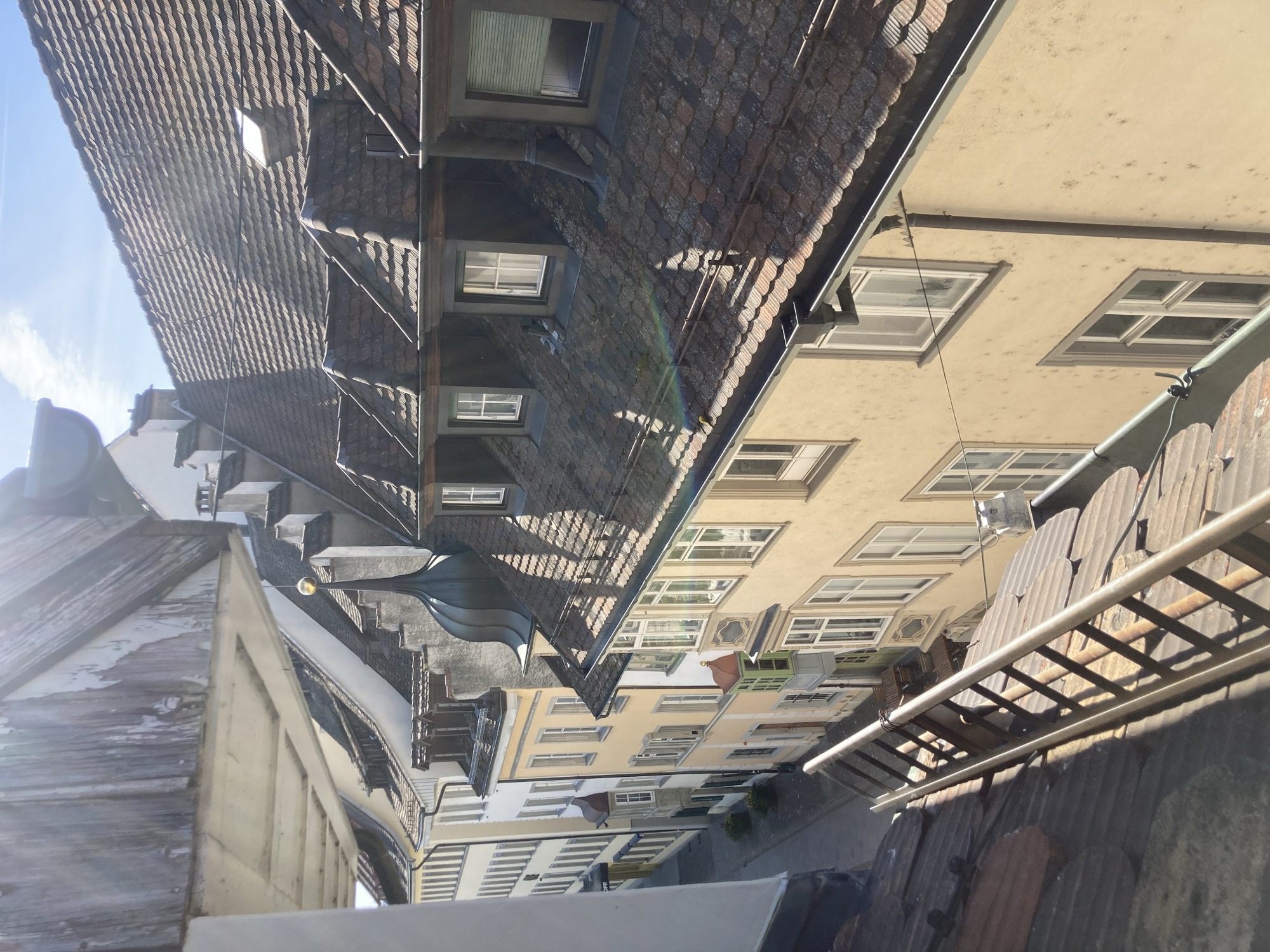 The image, originally rotated 90 degrees counter-clockwise, depicts a high-angle view looking down into a narrow street or alley lined with historic buildings in Schaffhausen, Switzerland. The scene is bathed in bright daylight, suggesting late morning or early afternoon.
Dominating the upper portion of the view are steeply pitched, dark tiled roofs, likely made of terracotta or slate, punctuated by numerous dormer windows. Below these, the building facades are predominantly light-colored, in shades of creamy yellow, beige, and white, featuring many rectangular windows with white frames. A distinctive building in the mid-ground boasts a dark, ornate spire or cupola. Another building exhibits a rustic, weathered wooden gable end.
In the foreground, a lower, dark tiled roof is visible, where a metal ladder rests horizontally. Utility lines or cables can be seen stretching between buildings. Direct sunlight, originating from the top-left (when corrected), casts sharp shadows and highlights the textures of the roofs and walls. A faint lens flare or rainbow effect is present in the sunlight. The visible sky is clear and blue with a few scattered clouds. The scene is devoid of human activity, conveying a sense of quietude in this historic European urban setting. No visible text is present.
