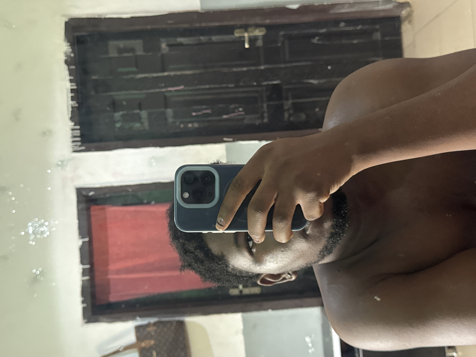 A dark-skinned person is taking a selfie in a mirror. The person is holding a blue smartphone with multiple rear-facing camera lenses. The background reflection shows a window with dark, wooden frames and what appears to be an interior space with red and brown objects. There is no information in the image to determine the location is Ipetumodu, Nigeria.