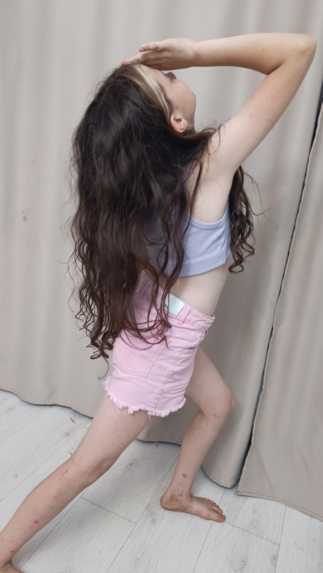 A young girl with long, wavy brown hair stands in a yoga pose. She is wearing a light purple crop top and light pink denim shorts with frayed edges. Her right arm is raised and bent, with her hand shielding her eyes as she looks up and to the right. Her left leg is extended forward in a lunge, and her right leg is bent behind her. The floor is made of light-colored wood planks, and a beige curtain hangs in the background. There are a few dark smudges on the girl's legs and feet, suggesting she may have been playing outdoors. The lighting is soft and diffused, indicating it is likely daytime indoors. The girl's pose and expression do not convey a specific emotion, but the overall impression is one of movement and flexibility. There is no visible text in the image. The location is Chaikovsky, Russia.