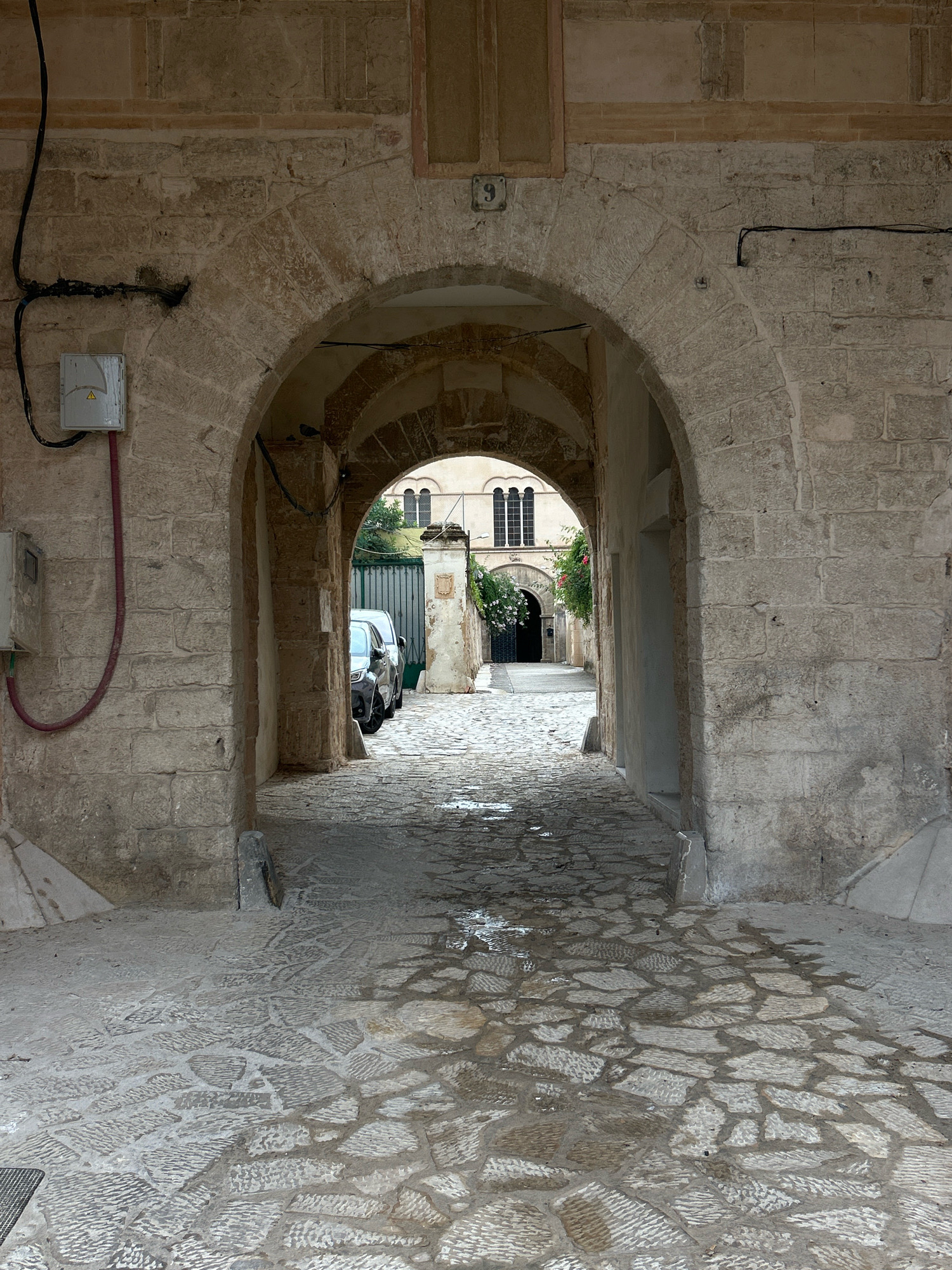 The image depicts an arched stone passageway in Palma, Spain. The passageway is constructed from rough-hewn, light-colored stone blocks. The ground is paved with irregular stone slabs that appear wet in places, reflecting the overhead arches.
On the left side of the passageway, a gray electrical box is mounted on the stone wall, with black cables running along the wall and up towards the ceiling. A red conduit snakes down from the box.
The passageway leads to an open courtyard or street area. Two cars are visible parked in the distance, partially obscured by the arches. Beyond the cars, a large green metal gate is visible, flanked by stone pillars. Above the gate, architectural details of a building can be seen, including a window with multiple panes and arched tops, and some greenery with red flowers trailing down.
The overall impression is of an old, possibly historic, urban environment in Palma, with a focus on the textures of the stone architecture and the wet pavement.
