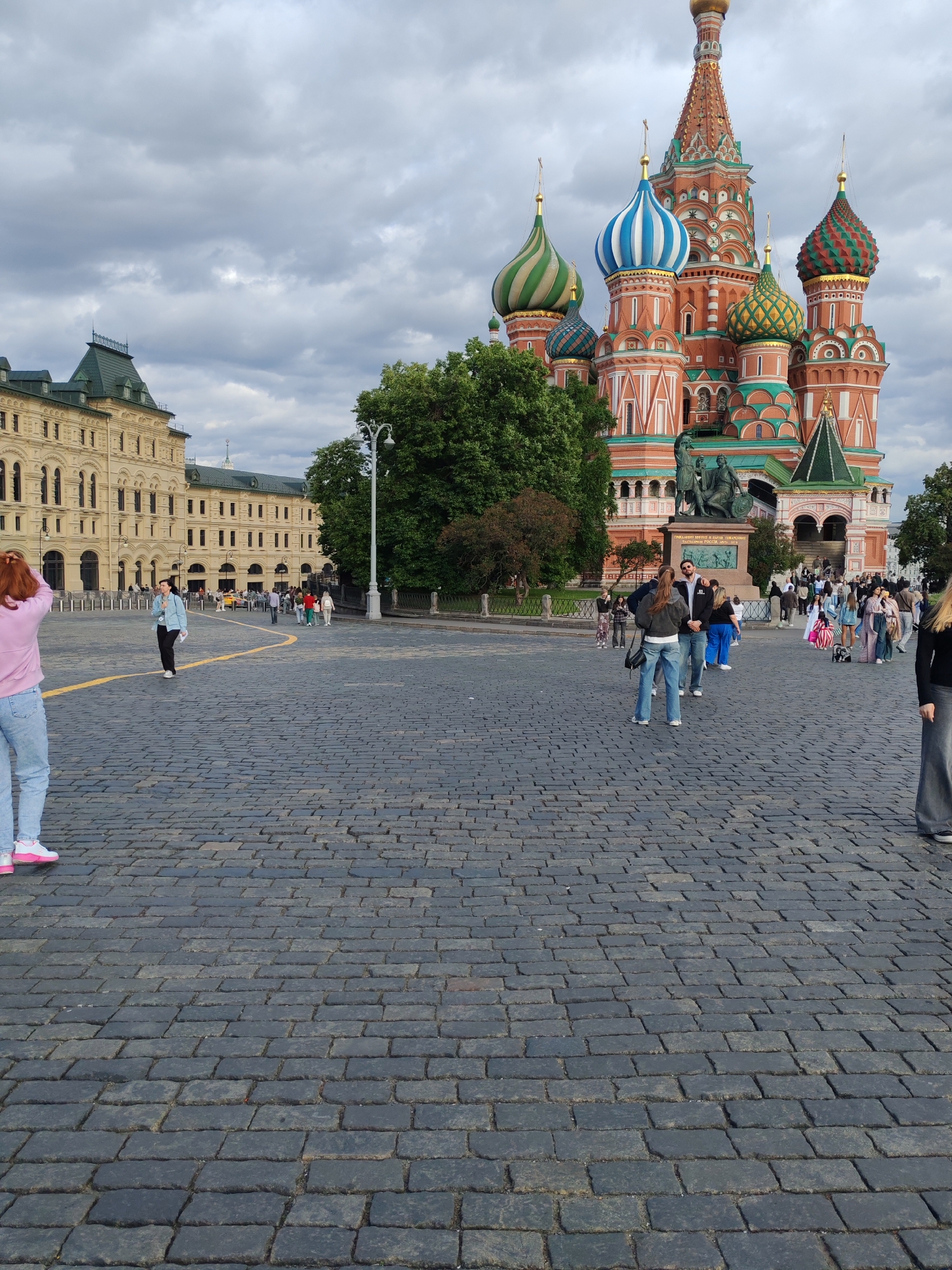In an outdoor urban setting with a cloudy sky, a paved plaza is filled with cobblestones. To the right of the frame, the iconic St. Basil's Cathedral, with its vibrant red and multi-colored onion domes, is a prominent landmark. In front of the cathedral, a bronze statue of figures on horseback stands on a pedestal. A crowd of people, dressed in casual attire, are scattered across the plaza, some walking, others standing in groups. To the left, a large, beige building with numerous windows and a green roof occupies a significant portion of the background. The scene captures a moment of everyday life in a public square.