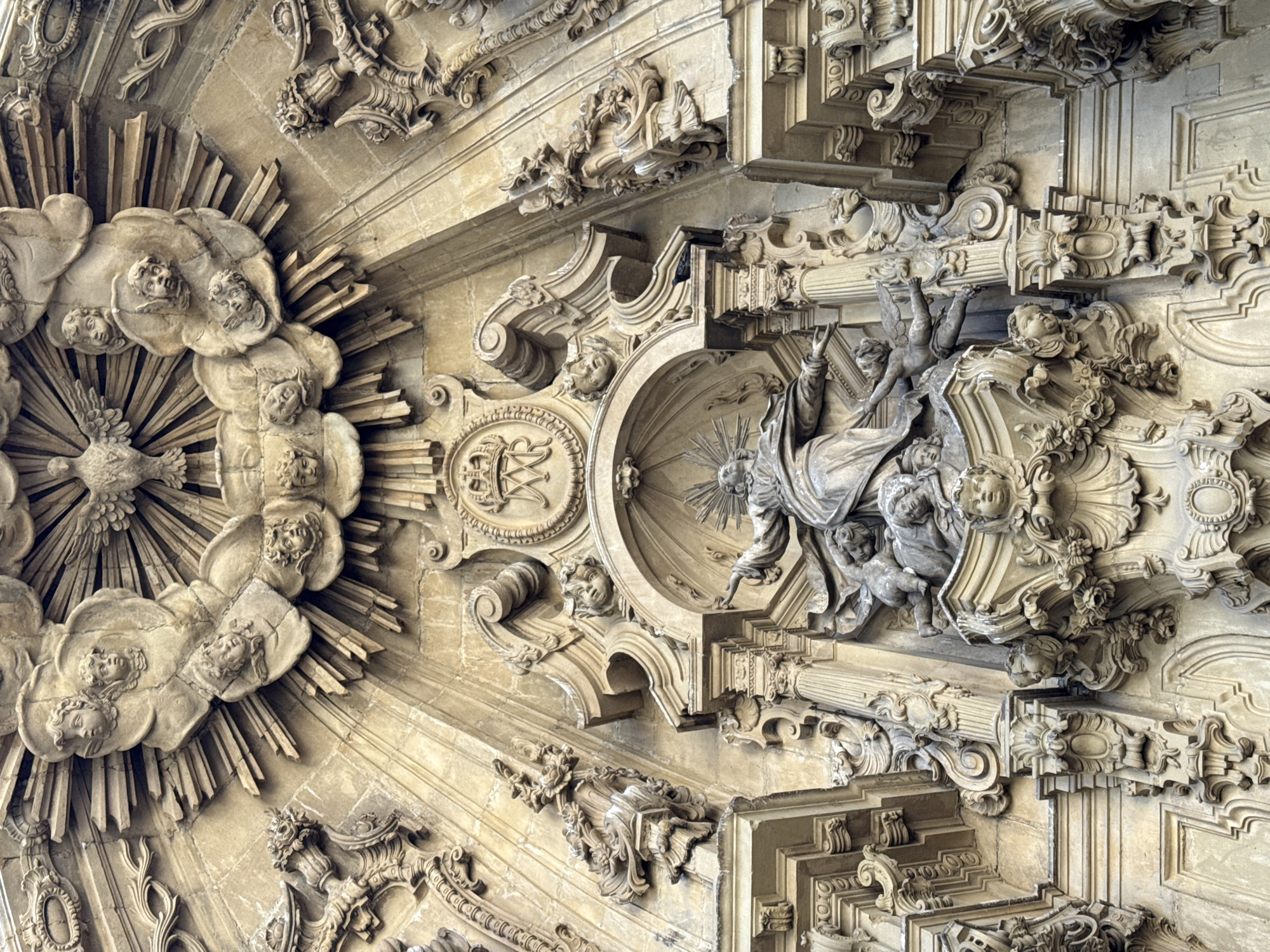 The image shows a close-up, low-angle view of the ornate facade of the Church of San Sebastián in San Sebastián, Spain. The sculpture is made of stone and depicts a central religious scene with a figure of a saint or divine being at the apex, surrounded by cherubic figures and a dove representing the Holy Spirit, all set against a backdrop of radiating rays. The detailed carvings extend down to include architectural elements like columns, decorative scrolls, and floral motifs. The stone appears weathered, indicating age and exposure to the elements.