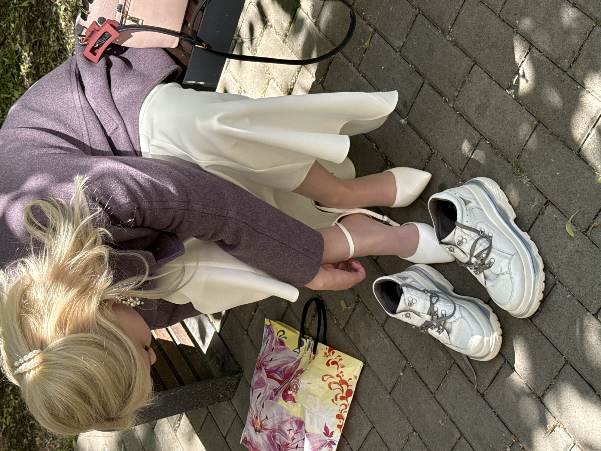 A blonde woman wearing a light-colored dress and a purple coat is sitting on a bench outdoors. She is in the process of changing her shoes. One white high-heeled shoe is on her foot, and the other is on the ground. Next to her are a pair of white sneakers. A floral-patterned shopping bag and a pink handbag are also visible. The scene takes place on a paved area with shadows from trees, suggesting it is daytime. The location is identified as Saint Petersburg, Russia.