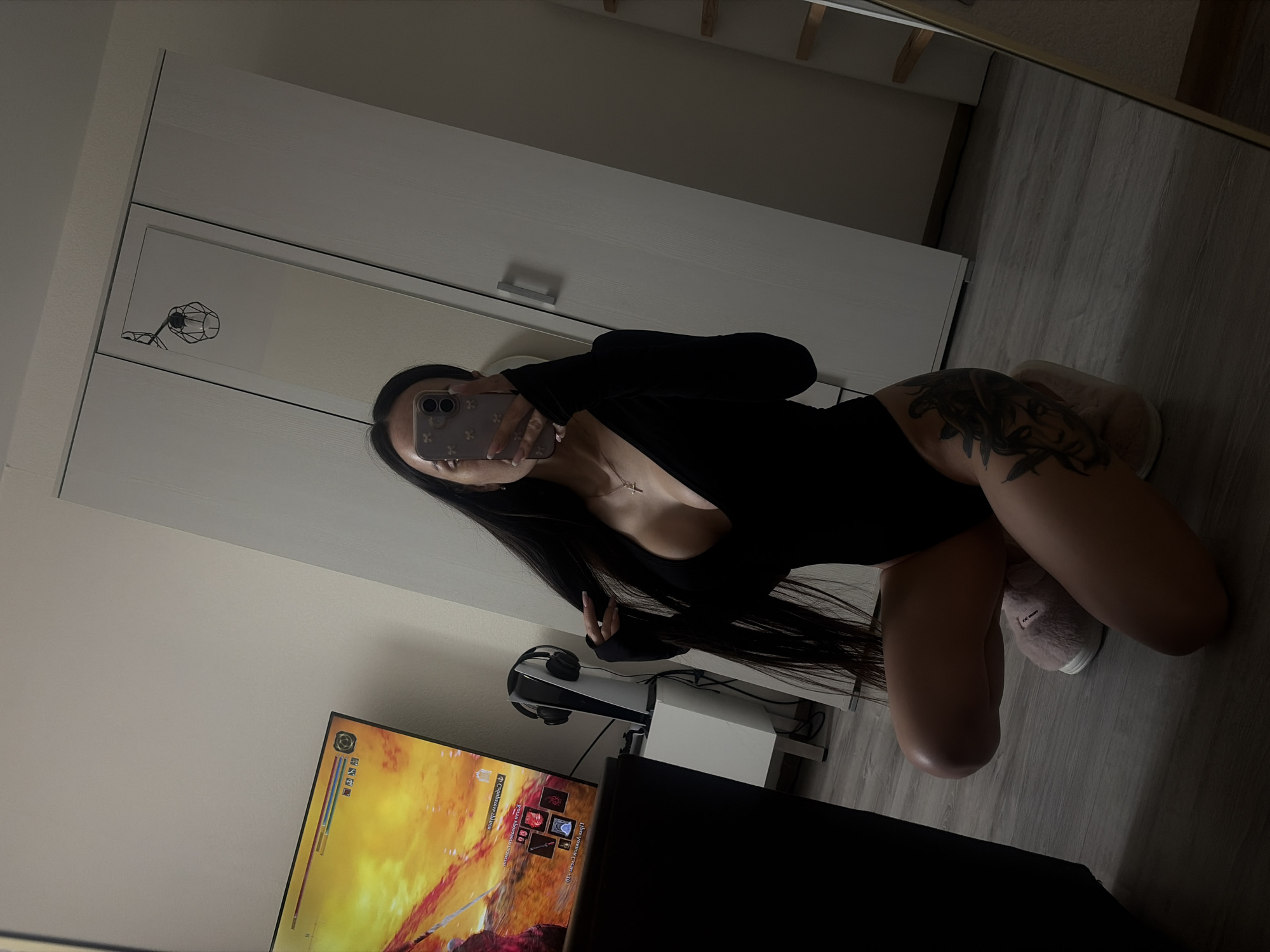 A dark-haired person is kneeling in front of a mirror, taking a selfie with a smartphone. The person is wearing a black, long-sleeved bodysuit and pink, fuzzy slippers. A tattoo is visible on the upper thigh. In the background, a television is displaying a video game, and headphones are placed on top of a white stand, which may be a gaming console. A white cabinet with two doors is also visible in the background. The location in the city of Saint Petersburg, Russia, cannot be determined from this image.