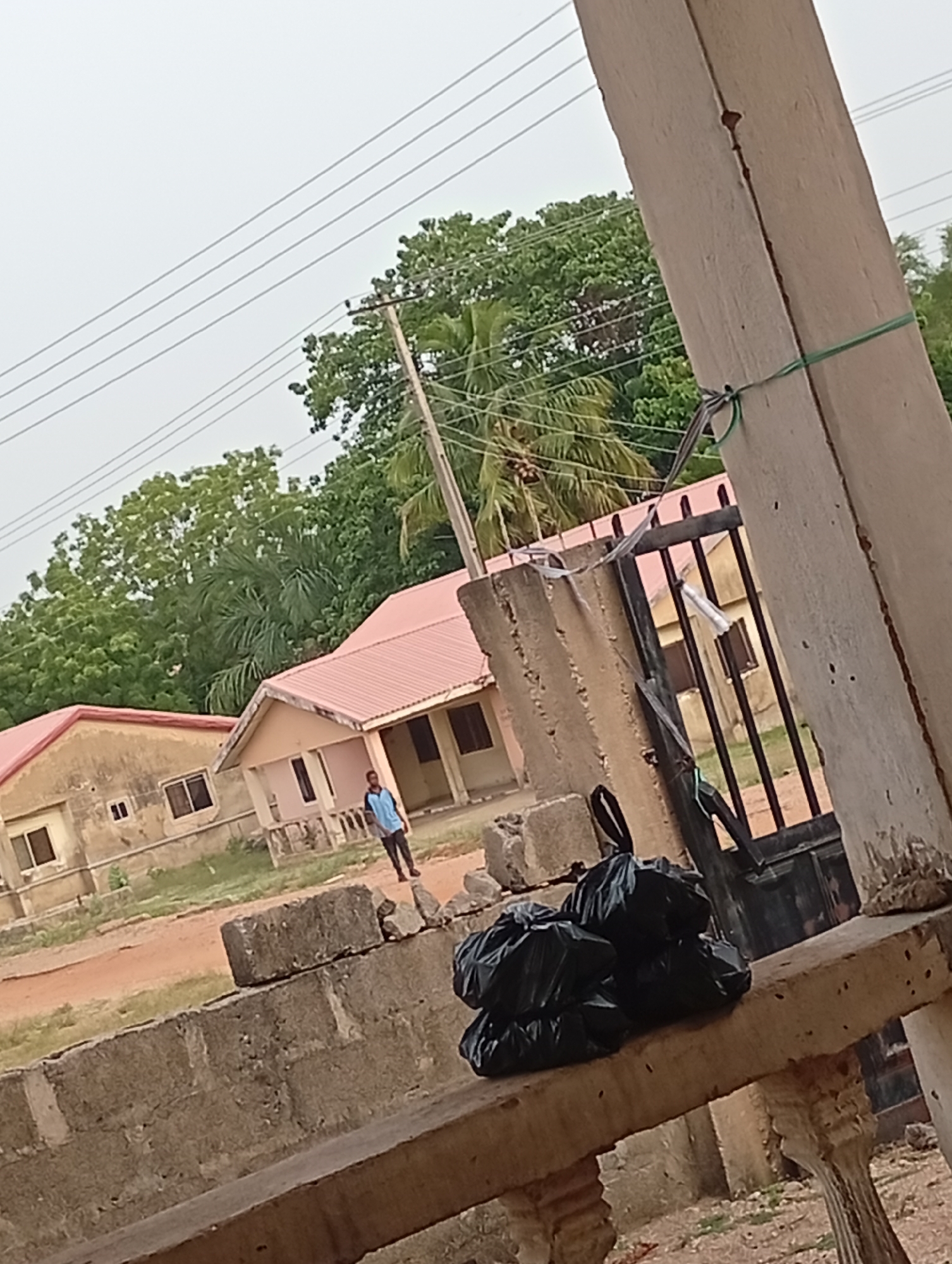 The image shows a partially obscured view of buildings and a person in an outdoor setting. In the foreground, there are several black trash bags on a concrete surface, with parts of a concrete pillar and a black metal gate visible. Beyond this, two buildings with pink roofs are seen, the one closer to the center of the frame appears to have a person standing in front of it. Trees and power lines are in the background. The image appears to have been taken in Unknown, Unknown.