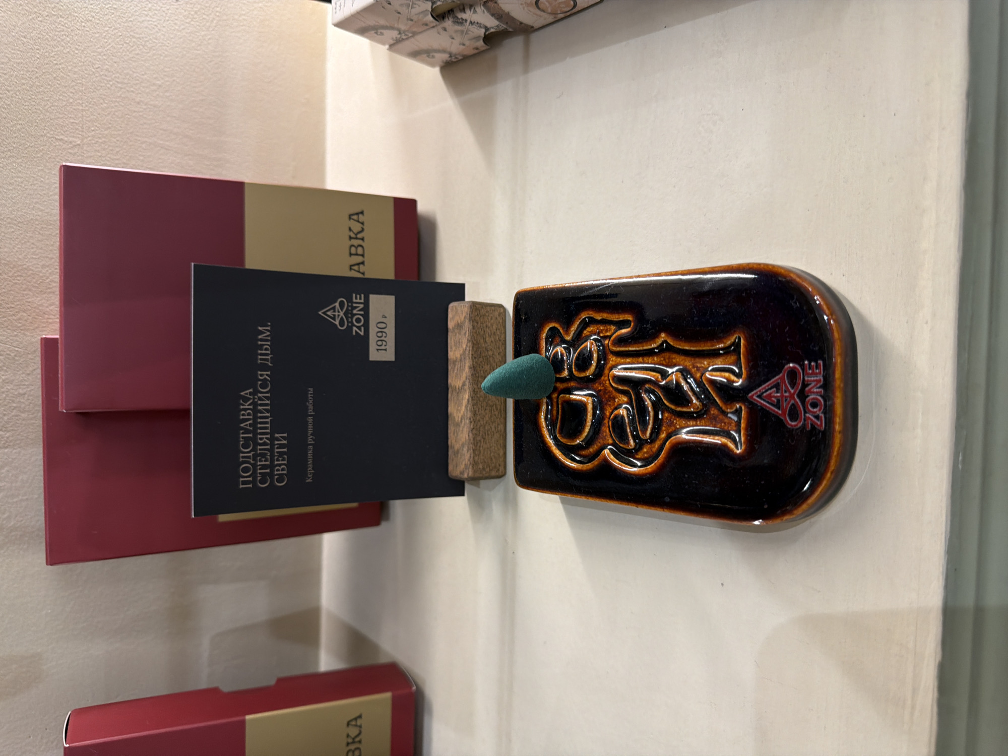 This image displays a product arrangement on a light-colored surface, likely a shelf in a retail environment. The orientation of the photo is portrait.
The central object is a glossy, dark brown ceramic incense burner, featuring raised, ornate designs in a lighter, orange-brown hue. A green, conical incense cone is positioned on a small, light-brown wooden block, which rests within the ceramic burner.
To the left of the incense burner, several product boxes are stacked. The most prominent is a dark blue box or tag with white text. Visible text on this item includes "ПОДСТАВКА СТЕЛЯЩИЙСЯ ДЫМ. СВЕТИ," which translates to "STAND FLOWING SMOKE. SHINE." Below this, "Керамика ручной работы" (Handmade ceramics) is written. A logo, depicting a triangular symbol next to the word "ZONE," is also present, along with a price tag showing "1990 Р" (1990 Rubles). Partially visible behind and below this blue item are several red or maroon boxes with lighter spines, one showing the partial text "АВКА." The "ZONE" logo is also visible in red on the ceramic incense holder itself.
The scene is a static product display, brightly lit indoors, with no people or dynamic activity occurring.