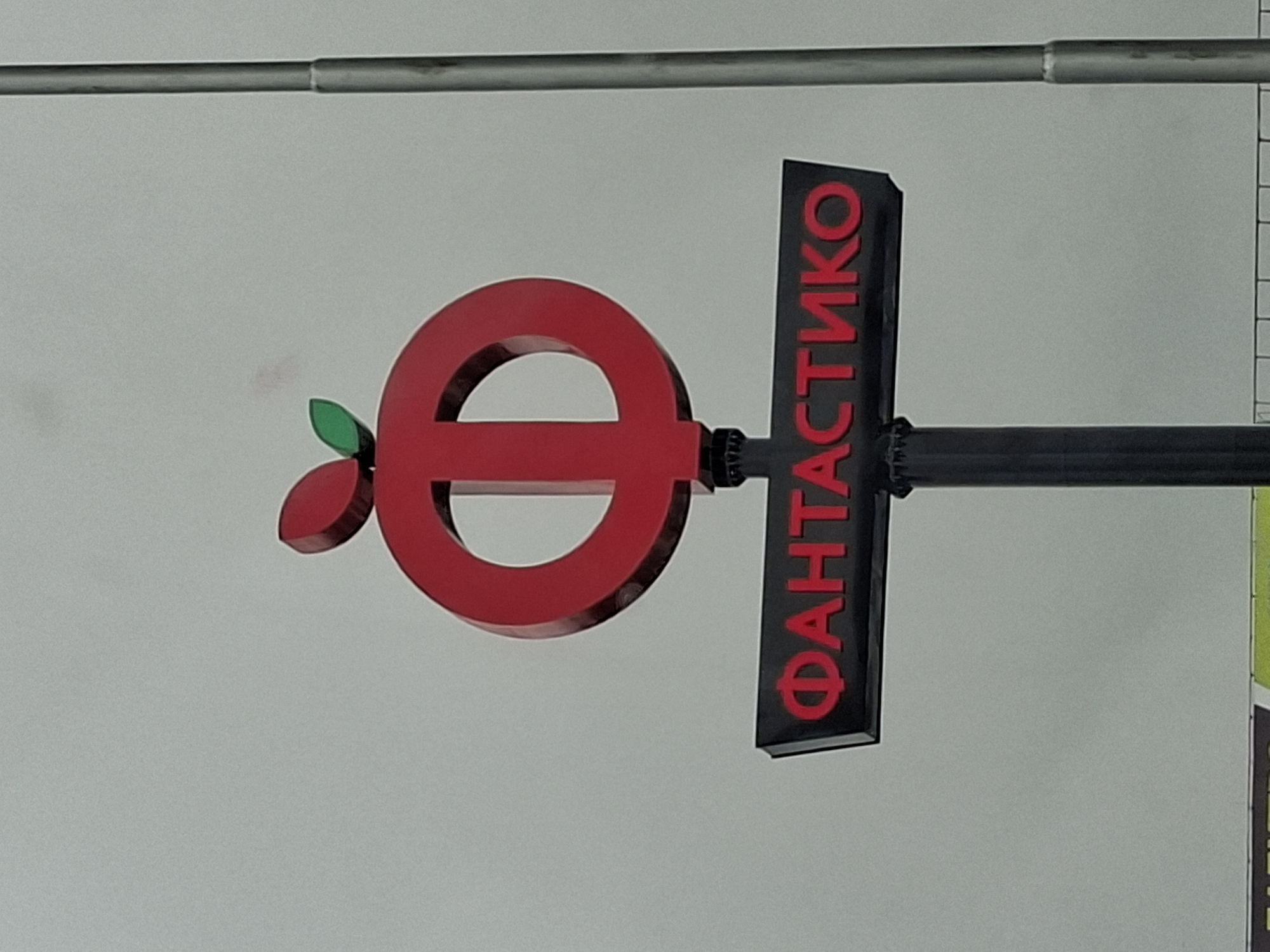 The image shows a sign for "ФАНТАСТИКО" (Fantastico) in Plovdiv, Bulgaria. The logo features a red circle with a horizontal bar through the middle, resembling a stylized apple with two green leaves. The text "ФАНТАСТИКО" is in red Cyrillic letters on a vertical black rectangle attached to the right side of the circle. The sign is mounted on a black metal pole. Above the sign is a horizontal metal bar. The background is a plain white wall. Part of another sign is visible on the right edge of the frame, and a measuring tape is present near the right edge of the frame.