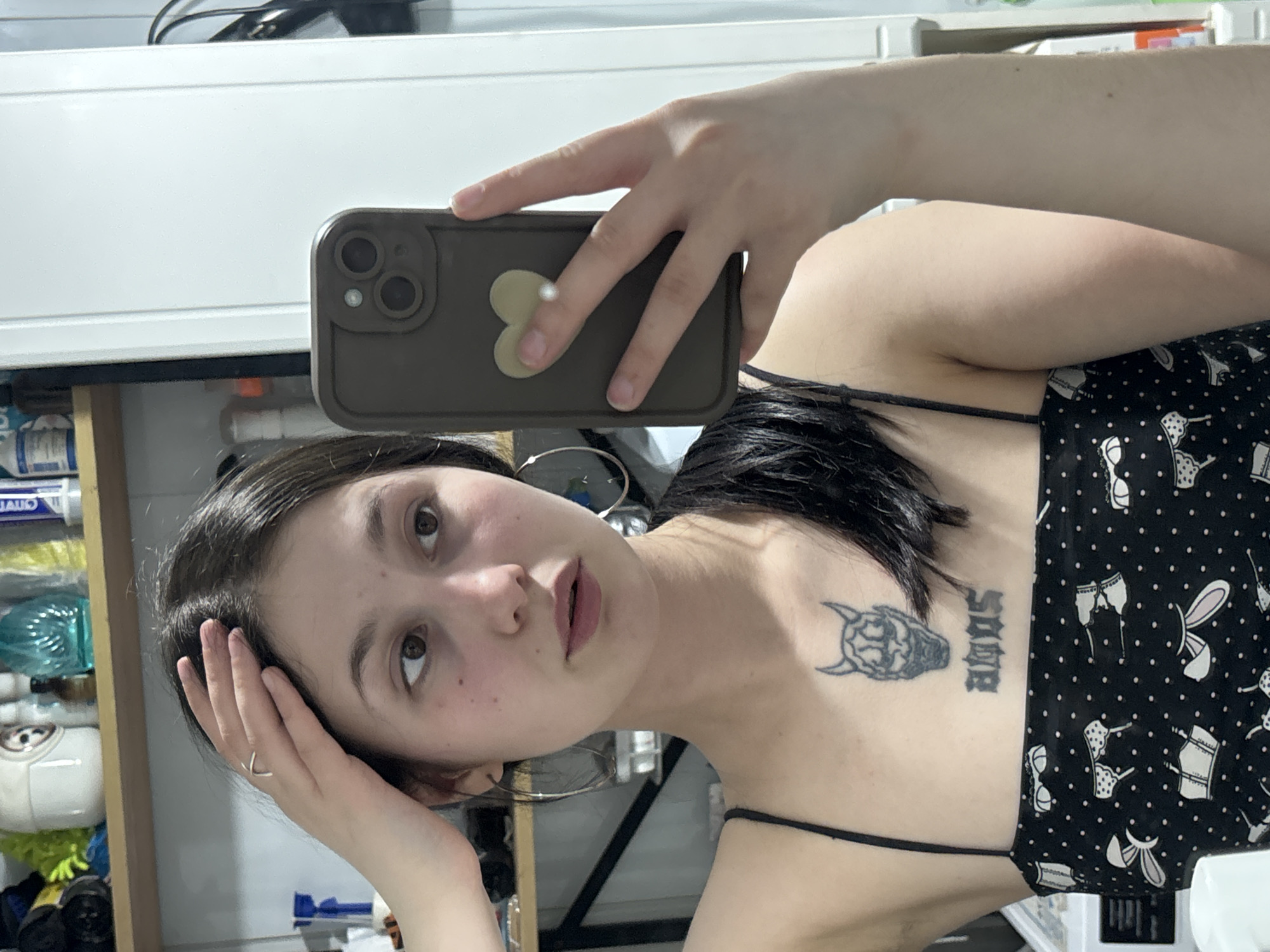 A young woman takes a selfie in an indoor setting. She is holding a smartphone with a light brown case, adorned with a heart-shaped sticker. She rests her head on her left hand, showcasing a simple silver ring. She has dark hair and brown eyes. She is wearing a spaghetti-strap black top with a white lingerie pattern. Tattoos are visible on her upper chest. The background includes a shelf with various items, a white wall or cabinet, and other indistinct objects. The location in the city of Фоки, Russia cannot be determined from the image.