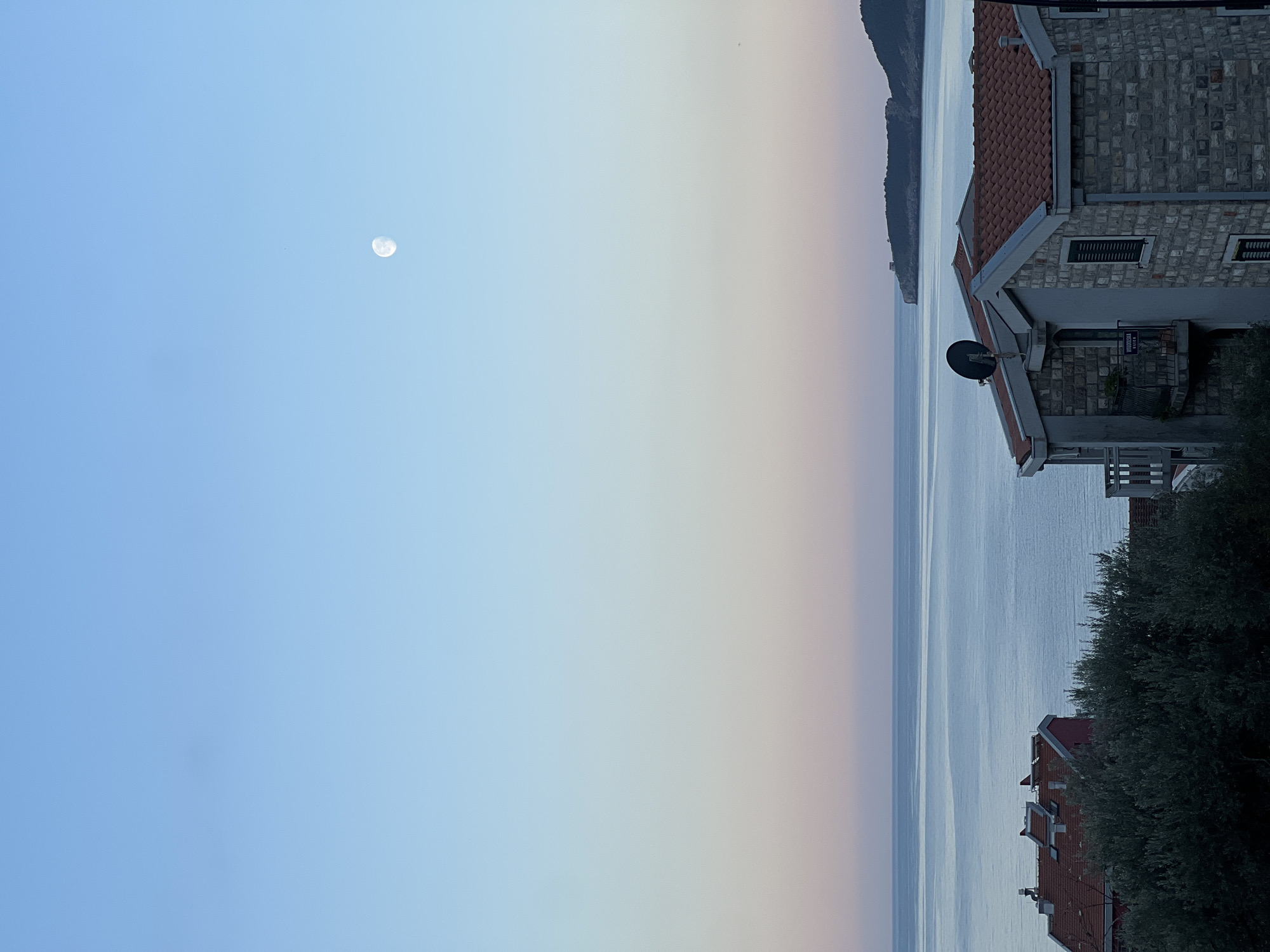 The image shows a seaside scene at dusk in Budva, Montenegro. A partial moon is visible in the light blue sky. Below the sky is a gradient to pale peach and then a calm sea. Buildings with red-tiled roofs and stone walls are on the right of the image, partially obscured by foliage. A peninsula is in the distance, extending into the sea.