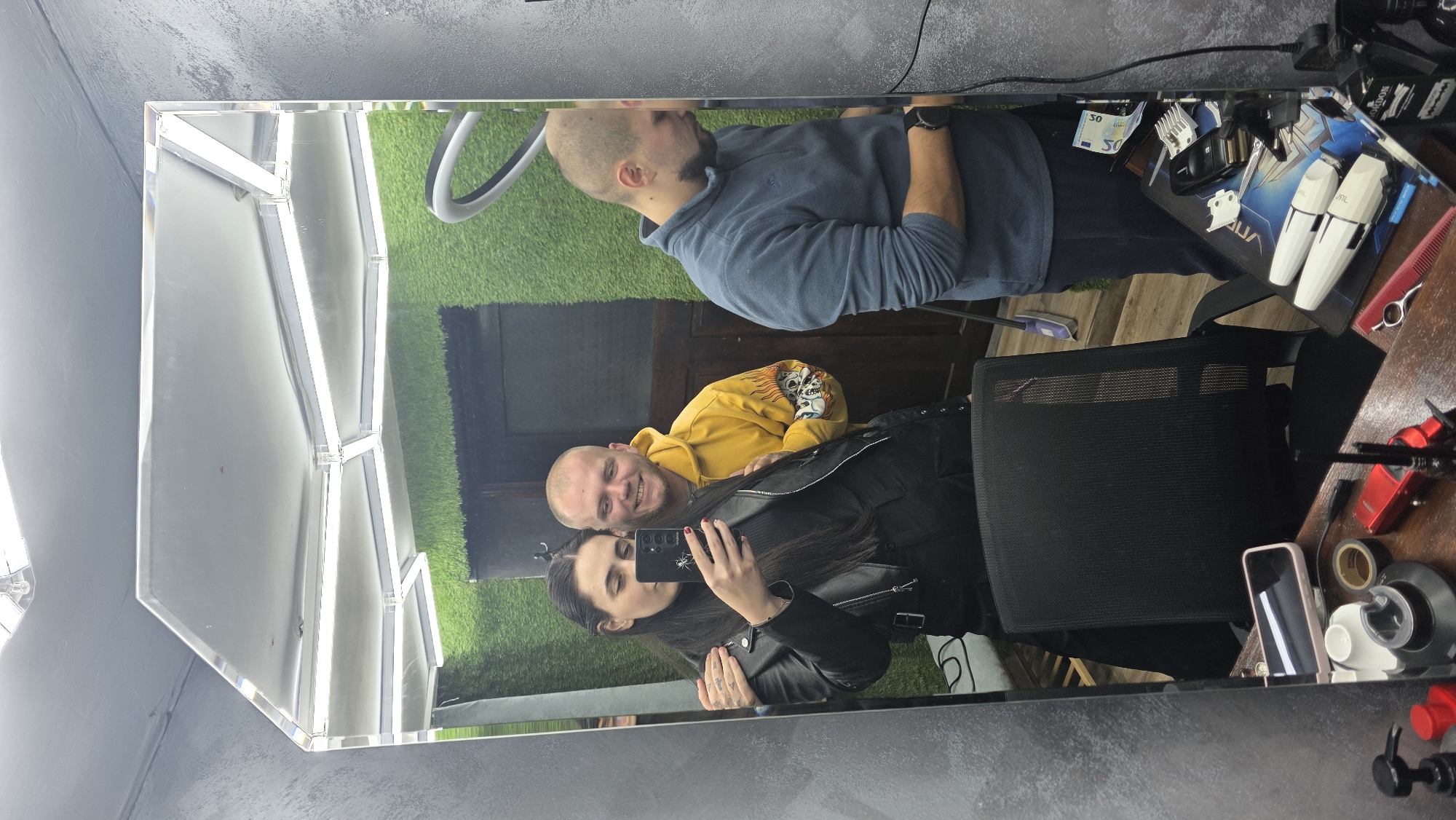 A mirrored image captures three individuals in what appears to be a barbershop or salon in Redina, Bulgaria. The primary subject is a woman with long dark hair, wearing a black leather jacket, who is holding a smartphone to take a selfie. She is smiling broadly and has tattoos visible on her hand. Standing behind her and also reflected in the mirror is a bald man with a beard, wearing a yellow hooded sweatshirt with a skull design, smiling towards the camera. Further in the background, another bald man with a beard, dressed in a blue long-sleeved shirt, is visible.
The setting includes a large mirror with integrated strip lighting along its edges. The background shows a decorative green artificial grass wall and wooden architectural elements. On a wooden counter to the right, various barber tools are laid out, including white and black hair clippers (one possibly branded "WAHL"), a red comb, scissors, and razors, all arranged on a black mat that appears to say "DUAL" or "AQUA." Two 50 Euro banknotes are also visible on the mat. A black mesh office chair is positioned in the foreground. Other items on the counter include a pink smartphone, a white cup, black tape, and some red objects. The overall mood conveyed by the smiles is cheerful and friendly.