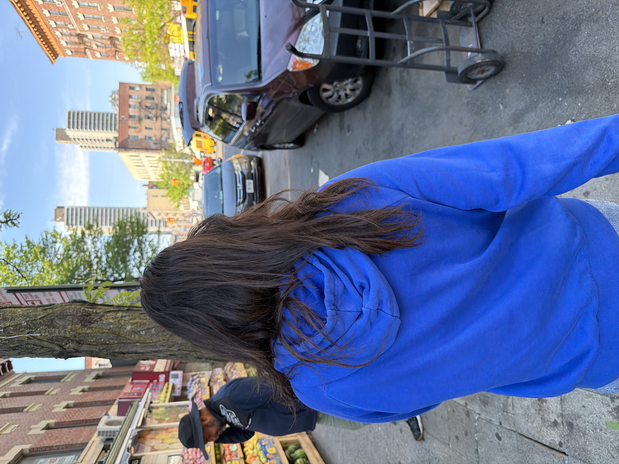 The image depicts an individual wearing a blue hooded sweatshirt standing on a city street. In the background, there are buildings typical of New York City, including both brick apartment buildings and taller, modern high-rises. Cars are parked along the street, and a hand truck is visible near one of the vehicles. Additionally, a fruit stand is present, with a vendor wearing a hat near the stand. The environment appears to be a bustling urban setting.