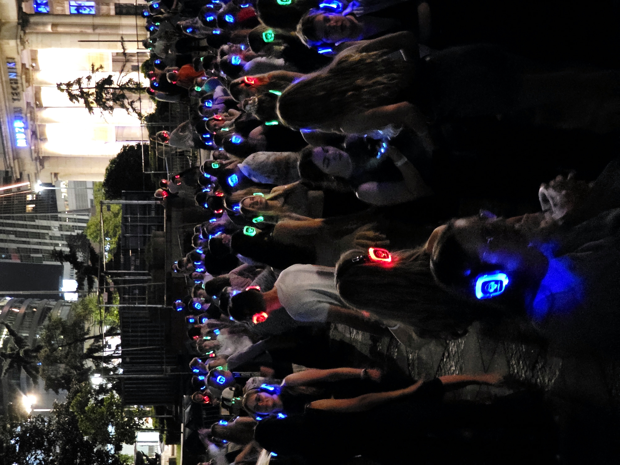 The image shows a large group of people wearing glowing headphones at night. The event appears to be a silent disco. The background features a building with the text "MUZEUM TECHNIKI" visible, indicating the location is the Museum of Technology in Warsaw, Poland. The people are diverse in age and gender, and they are standing and interacting in a relaxed, informal setting. The headphones are illuminated with various colors, including blue, red, and green, creating a vibrant and dynamic visual effect. The overall atmosphere is festive and energetic, suggesting a fun and engaging experience for the attendees.