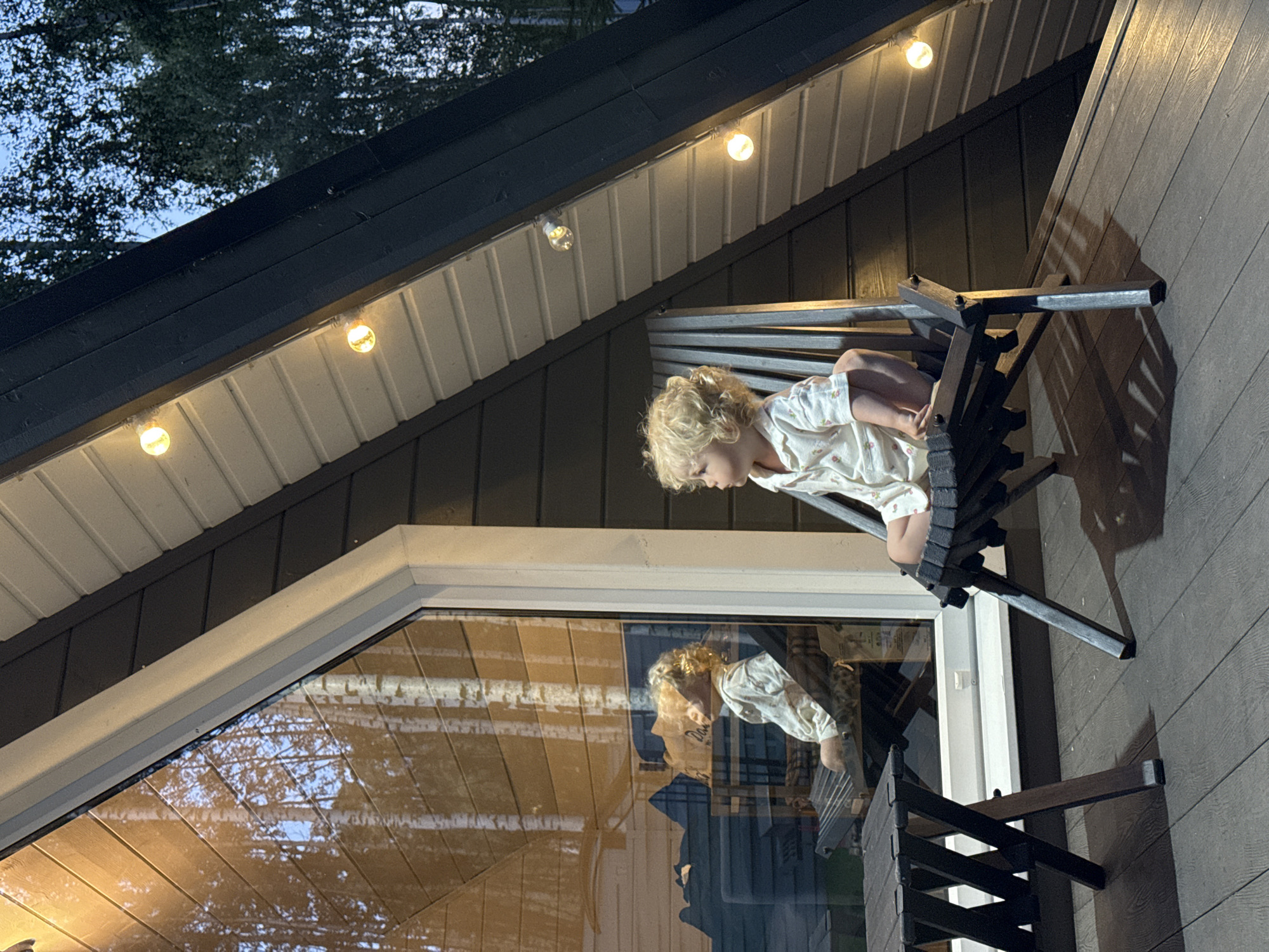 A young child with curly blonde hair sits on a dark wooden folding chair on a wooden deck. The child is wearing a light-colored pajama set with a floral print. The child is looking to the left, with their reflection visible in a large window. The window reflects trees and the interior of a building. The building has dark brown siding and a white-trimmed triangular roofline with string lights. A dark wooden folding table is situated to the left of the child. The scene is set in Prokudino, Russia, likely in a residential area with surrounding trees. The time of day appears to be dusk or early evening, as indicated by the ambient light and the glow of the string lights.
