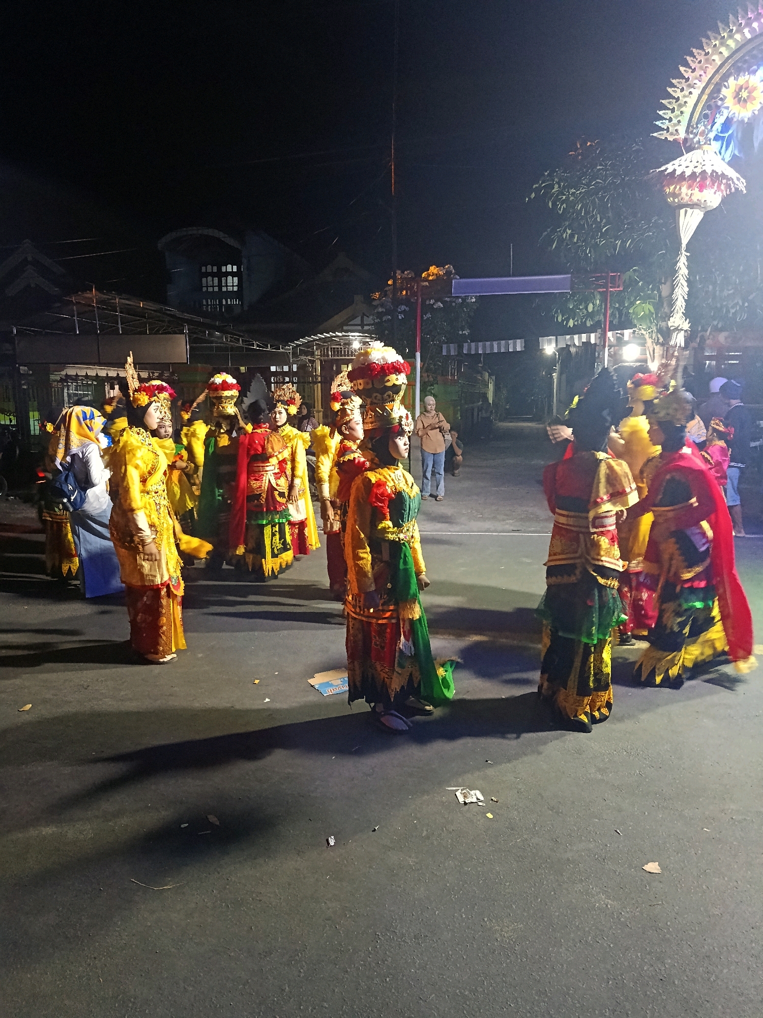 The image depicts a nighttime street scene in Pasirian, Indonesia, likely during a cultural event or parade. Several individuals, primarily women, are dressed in vibrant, traditional attire characterized by elaborate headdresses, colorful fabrics, and ornate skirts. The clothing appears to be ceremonial. Some individuals are standing, while others seem to be in motion, suggesting a procession or performance. In the background, a building with windows is visible, along with trees and a decorated archway, indicating an urban or semi-urban setting. The lighting is artificial, with streetlights and possibly lighting from the event itself illuminating the scene. Shadows are cast on the asphalt ground, emphasizing the nighttime setting. The overall atmosphere suggests a festive and culturally significant occasion.