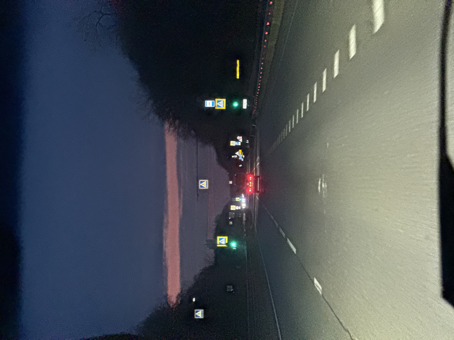 The image displays a multi-lane asphalt road in Khanskaya, Russia, at twilight or nightfall. A red car with illuminated tail lights proceeds in the distance, centered on the roadway. The road surface features white dashed and solid lane markings, with the foreground illuminated by headlights. A green traffic signal is active on the left side of the road, accompanied by multiple illuminated yellow pedestrian crossing signs. On the right, a vertical sign stack shows a blue bus stop icon above a yellow pedestrian crossing sign. A guardrail with red reflective elements lines the far right shoulder. Dense, dark vegetation borders both sides of the road. The sky exhibits a dark blue hue with a prominent, horizontal strip of pink light near the horizon. No individuals are present.