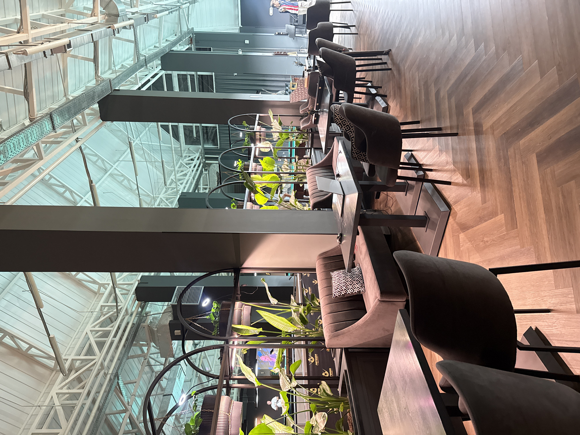 The image depicts the interior of a modern cafe or restaurant with a high industrial-style ceiling. The space features dark gray pillars, a herringbone patterned wooden floor, and various seating arrangements. In the foreground, there are dark gray tables with matching chairs. To the left, a U-shaped, plush gray sofa with a geometric patterned pillow is visible, situated next to a dark table with a laptop. Behind the sofa, a large, dark-colored shelving unit filled with lush green plants and illuminated by spotlights adds a decorative element. Further back, more seating and tables are arranged, with some individuals visible in the background, including two men near a pool table. The overall ambiance is contemporary and spacious, with a focus on natural light filtering through the high ceiling and artificial lighting from the plant displays and overhead fixtures. The location is identified as Balaklavsky District, Ukraine.