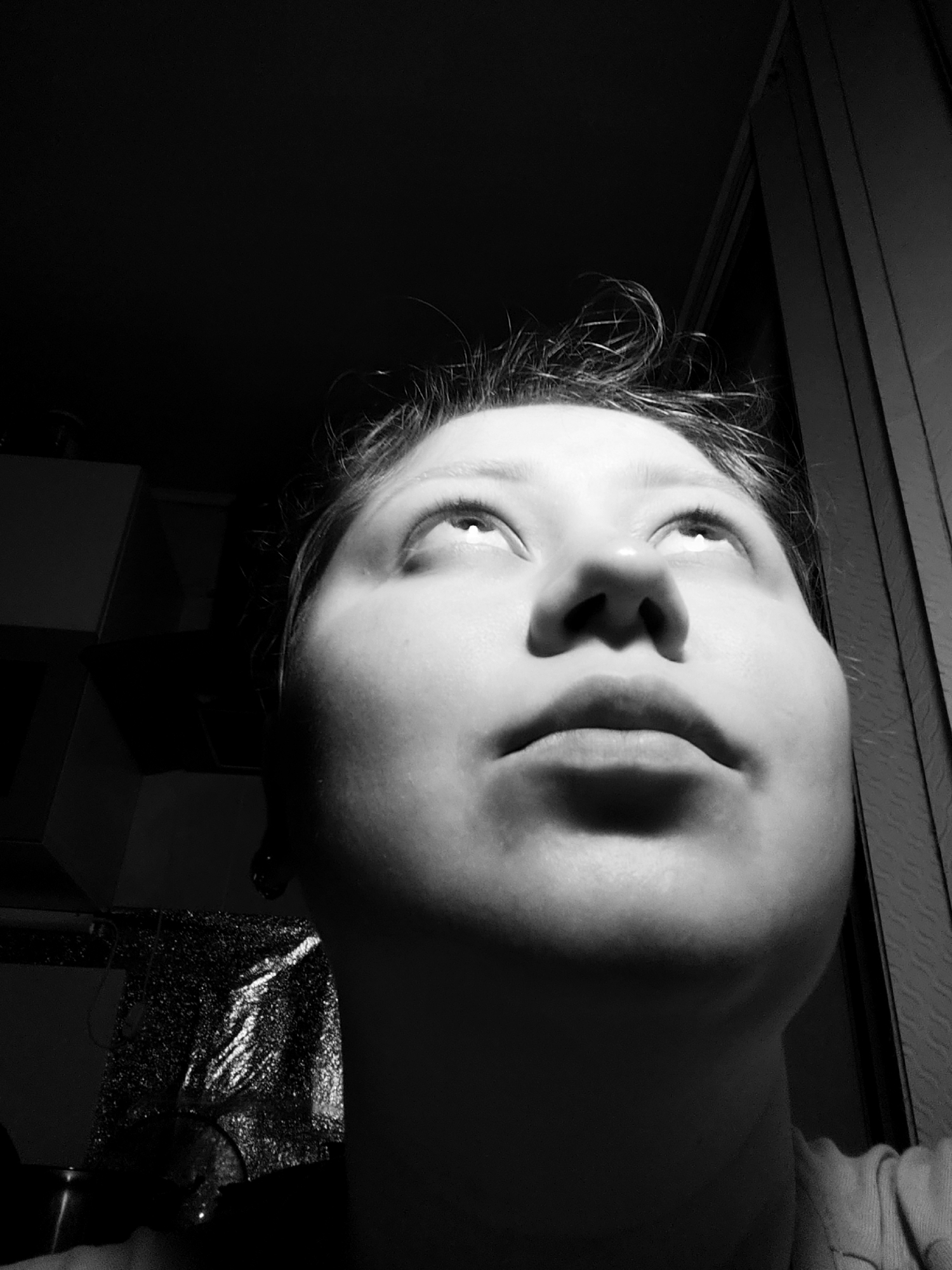 A low-angle, black and white photograph depicts a close-up of a young person's face looking upwards, in Fergana City, Uzbekistan. The lighting is dramatic, with a strong light source coming from above and to the left, casting deep shadows across the face and obscuring much of the background. The subject's eyes are open and looking upwards, with highlights visible in their irises. Their mouth is slightly open. The person has dark, messy hair that is styled upwards.
The background is mostly dark, but to the left, the edge of a light-colored object, possibly a piece of furniture or a wall, is visible. To the right, a textured surface, likely a curtain or blinds, runs vertically along the edge of the frame. The lower left of the image shows a glinting, textured surface that could be crinkled foil or a metallic fabric. The overall impression is one of introspection or contemplation.