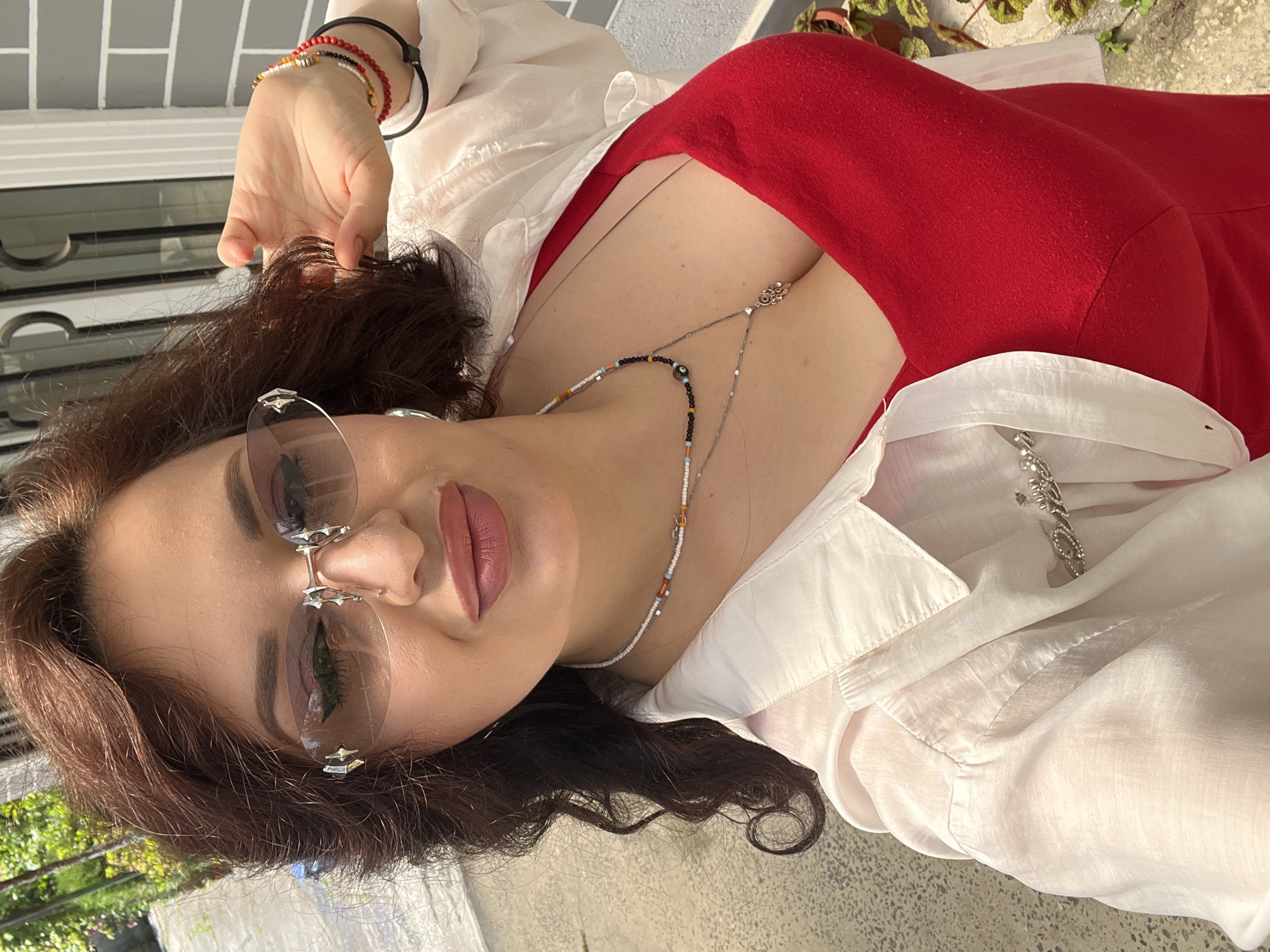 A woman poses for a selfie outdoors. She has auburn hair styled in loose curls, wears large rimless sunglasses, and has on a red dress and an open white collared shirt over it. She is accessorized with a beaded necklace and several bracelets. Her left hand is raised to her hair. The background shows a grey tiled wall and some greenery.