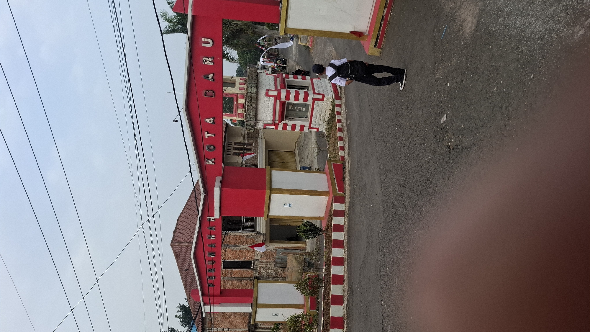 The image shows an entrance archway to a residential area in Cilaku, Indonesia, labeled "PERUMAHAN KOTA BARU". The archway is painted red and white, with "PERUMAHAN" on the left side and "KOTA BARU" on the right. A young person wearing a white shirt, dark pants, a black backpack, and a black cap stands facing away from the camera, looking towards the archway. The person is on the asphalt road in front of the entrance. The environment is a street with some buildings behind the archway, including one with exposed brickwork and another with a garage door. There are also some small trees and decorative elements along the roadside. The sky is overcast.