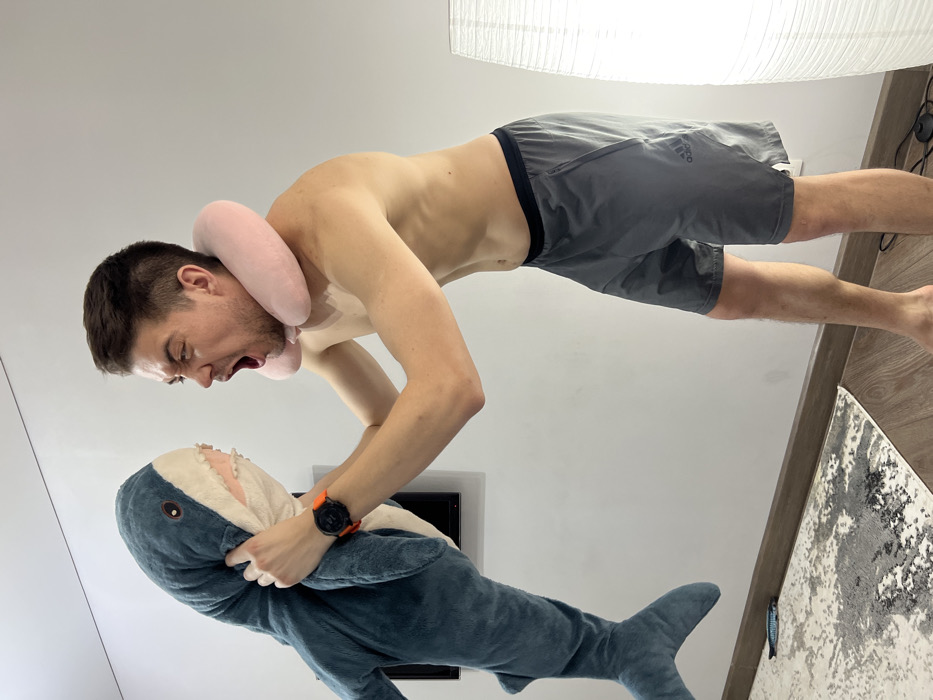 The scene depicts an adult male indoors, engaged in a confrontational pose with a large plush shark toy. The male, shirtless and barefoot, wears grey Adidas shorts and a light pink U-shaped neck pillow. An orange-strapped sports watch is visible on his left wrist, and a light-colored medical patch is present on his upper right chest. He is holding the blue-grey plush shark by its head with both hands. His mouth is wide open, emitting a scream or roar, with eyes squinted and teeth visible, directly facing the open mouth of the shark. The background consists of plain white walls, light wood-toned flooring with a dark baseboard, and a grey and white abstract patterned rug. A wall-mounted flat-screen television or monitor is visible behind the shark, and a tall, white pleated floor lamp stands in the right foreground. A small, blue-patterned object resembling a toy fish lies on the rug to the left. The environment is an interior space, specifically a residence in Kazan, Russia.