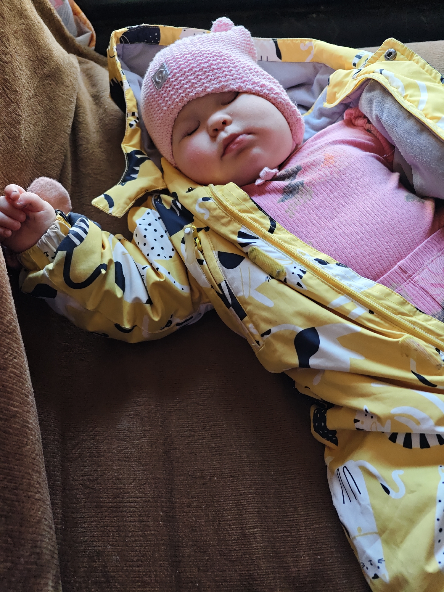 A baby wearing a pink knit hat and pink pajamas is sleeping on a brown couch while partially covered by a yellow patterned jacket. The jacket has a design of black and white abstract animal shapes. The couch is brown and textured. The location is presumed to be inside a home or dwelling, based on the presence of furniture, but nothing in the image directly indicates this is Malyi Bukor, Russia.