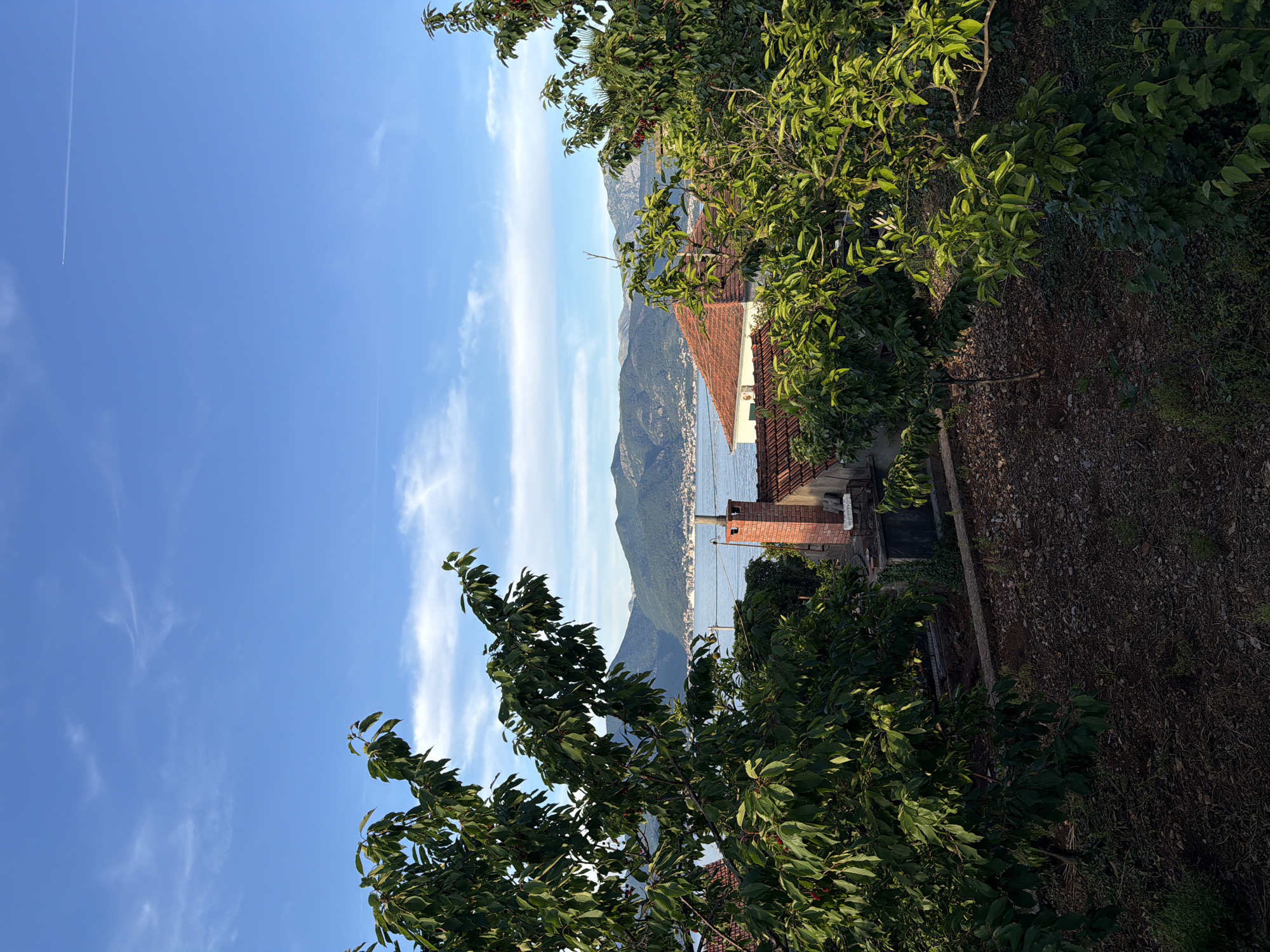 A slightly off-vertical, daytime shot presents a view in Krašići, Montenegro, with a clear, blue sky dominating the scene, marked by wispy clouds and a contrail from an aircraft.
The foreground is filled with lush green trees. In the midground, a red-tiled roof of a building is visible, accompanied by a brick chimney. Further in the distance, across a body of water, lies a mountain range. The landscape suggests a setting where residential areas blend into the natural terrain.