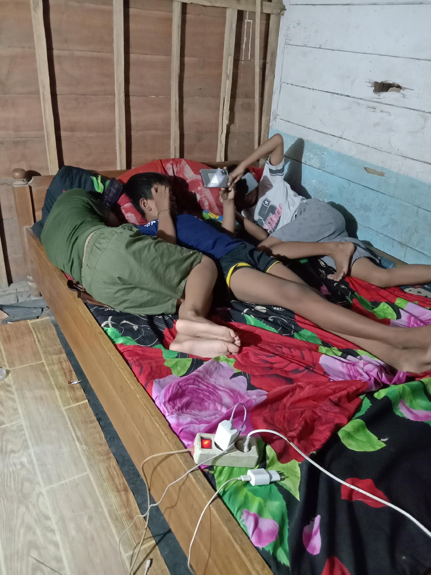 In a dimly lit room, likely in Jumput, Indonesia, three children are lying on a bed with a floral-patterned bedsheet. The child on the far left is asleep, facing away from the camera, covered by a green garment. The middle child is also asleep, with their face partially obscured by their hand. The child on the right is awake and holding a smartphone, appearing to be engrossed in its screen. The room's walls are constructed of rough wooden planks, with one section painted a faded blue. A power strip with two white charging adapters is plugged into the wall near the bed, with white cords trailing across the bedding. The floor appears to be wooden.
