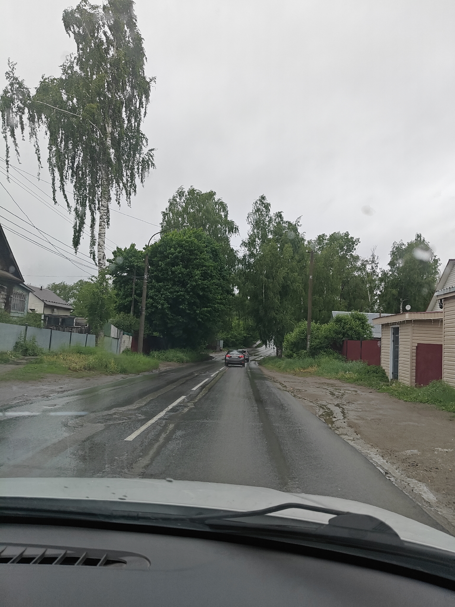 The image depicts a wet road in a residential area, likely in Chaikovsky, Russia, given the style of architecture. The view is from inside a car, looking down the street. A car is ahead. Trees line both sides of the road. The weather appears overcast or rainy. Buildings, fences, and power lines are visible on either side of the street.