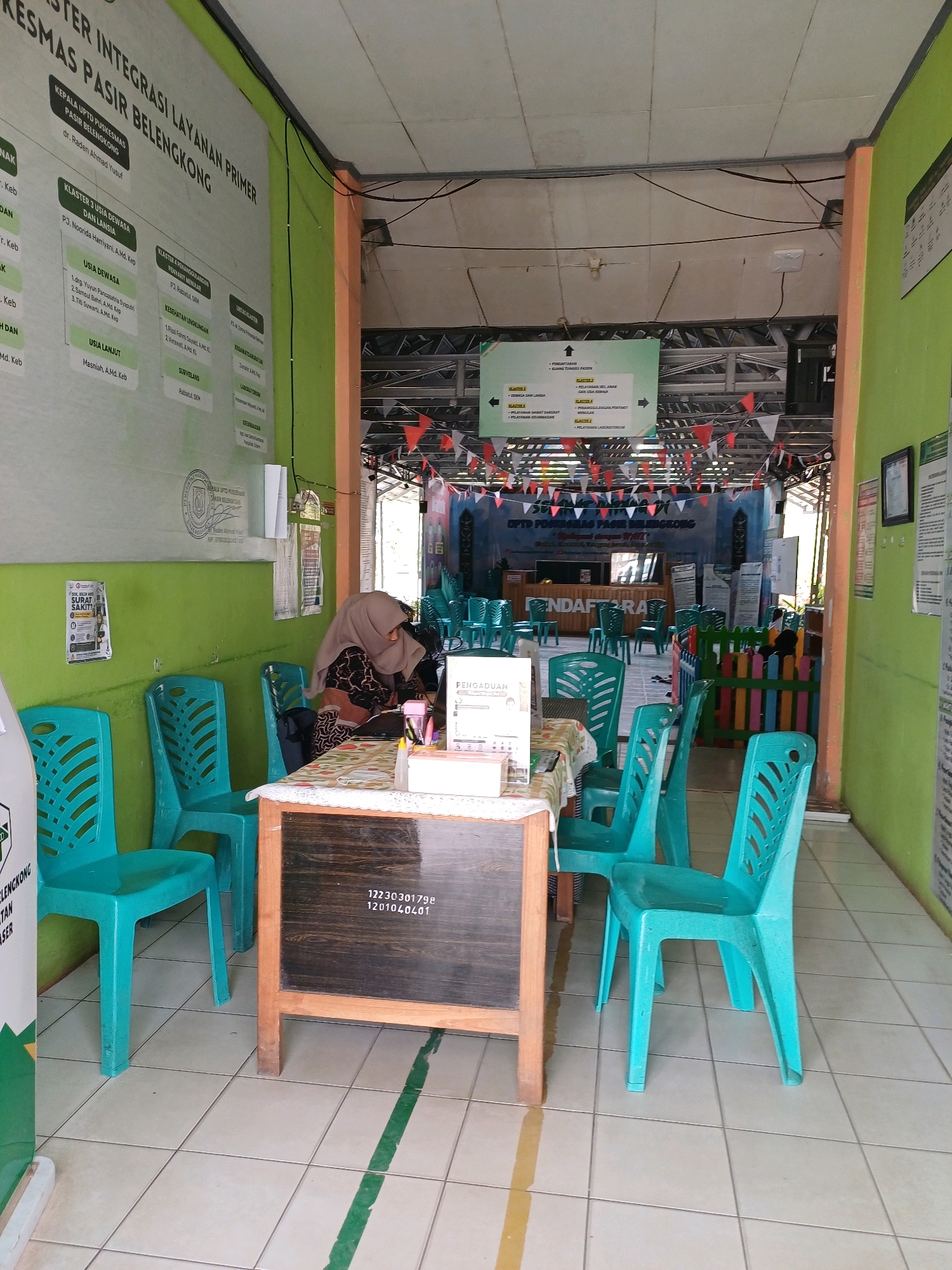 The image depicts the interior of a health center in Pasir Belengkong, Indonesia, likely a Puskesmas or similar public health facility. The walls are painted a bright green and orange. In the foreground, several teal plastic chairs are arranged. A woman wearing a hijab and a patterned dress is seated at a table, appearing to be working. On the table, there are papers, a pink bottle, and other office supplies. A small chalkboard with some numbers written on it stands in front of the table. Behind the woman, a sign with organizational charts and information is displayed on the wall. Further back, more teal chairs are visible, and a decorated area with a banner that reads "PENDAFTARAN" (Registration) is set up, suggesting a waiting or registration area. Festive triangular flags are strung across the ceiling. The floor is tiled. The overall impression is of a functional, if somewhat basic, public service environment.