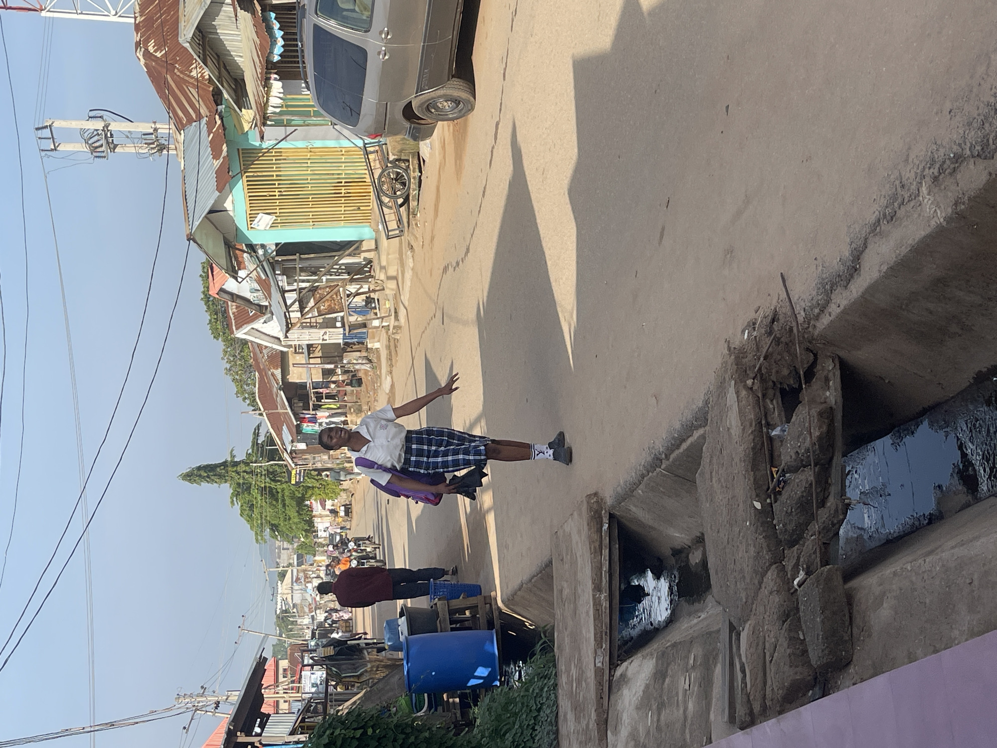 This image captures a bright, sunny street scene in what appears to be an urban or semi-urban area in Abuja, Nigeria.
The main subject is a young person, likely a student, positioned in the middle of the unpaved street. They are wearing a white short-sleeved collared shirt, a blue and white plaid skirt, white socks with black stripes, and dark shoes. A purple backpack is visible on their left shoulder, and they hold a black item (possibly an umbrella or bag) in their left hand. Their right arm is slightly extended, and they look towards the camera with a neutral expression.
To the right of the street, a large, open concrete drainage ditch runs parallel, containing dark, stagnant water and some debris. On the left side of the street, two large blue plastic barrels are visible, along with a maroon-clad person facing away from the camera, and another individual behind them. Further down the street, various people and vehicles, including motorcycles, are present.
Buildings with corrugated metal roofs line both sides of the street. Some structures feature light blue walls and yellow security grates. A grey minivan is parked on the left, with a metal pushcart nearby. A tall wooden utility pole with multiple power lines stands prominently on the left side of the road. In the background, green trees are visible against a clear, blue sky.
The strong shadows cast on the ground suggest it is either morning or early afternoon on a clear, sunny day. The word "VELLA" might be discernible vertically on the side of the grey minivan.