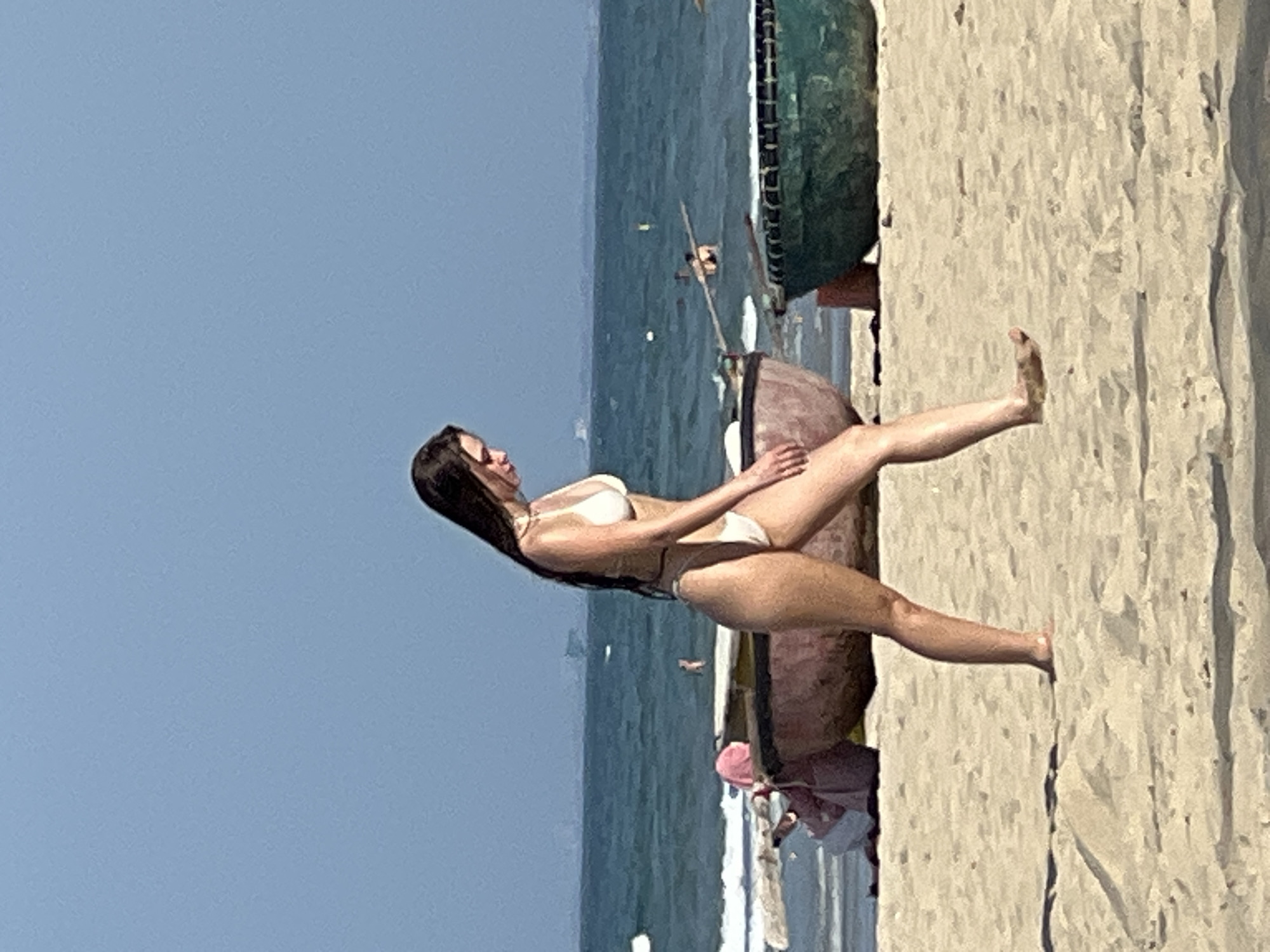 A woman in a white bikini walks on a sandy beach with the ocean in the background. She has long dark hair and is looking down. Two round basket boats are on the sand behind her, one pinkish-brown and the other green. In the distance, the ocean is calm with a hazy blue sky above. The scene is consistent with a beach in Đà Nẵng, Vietnam.