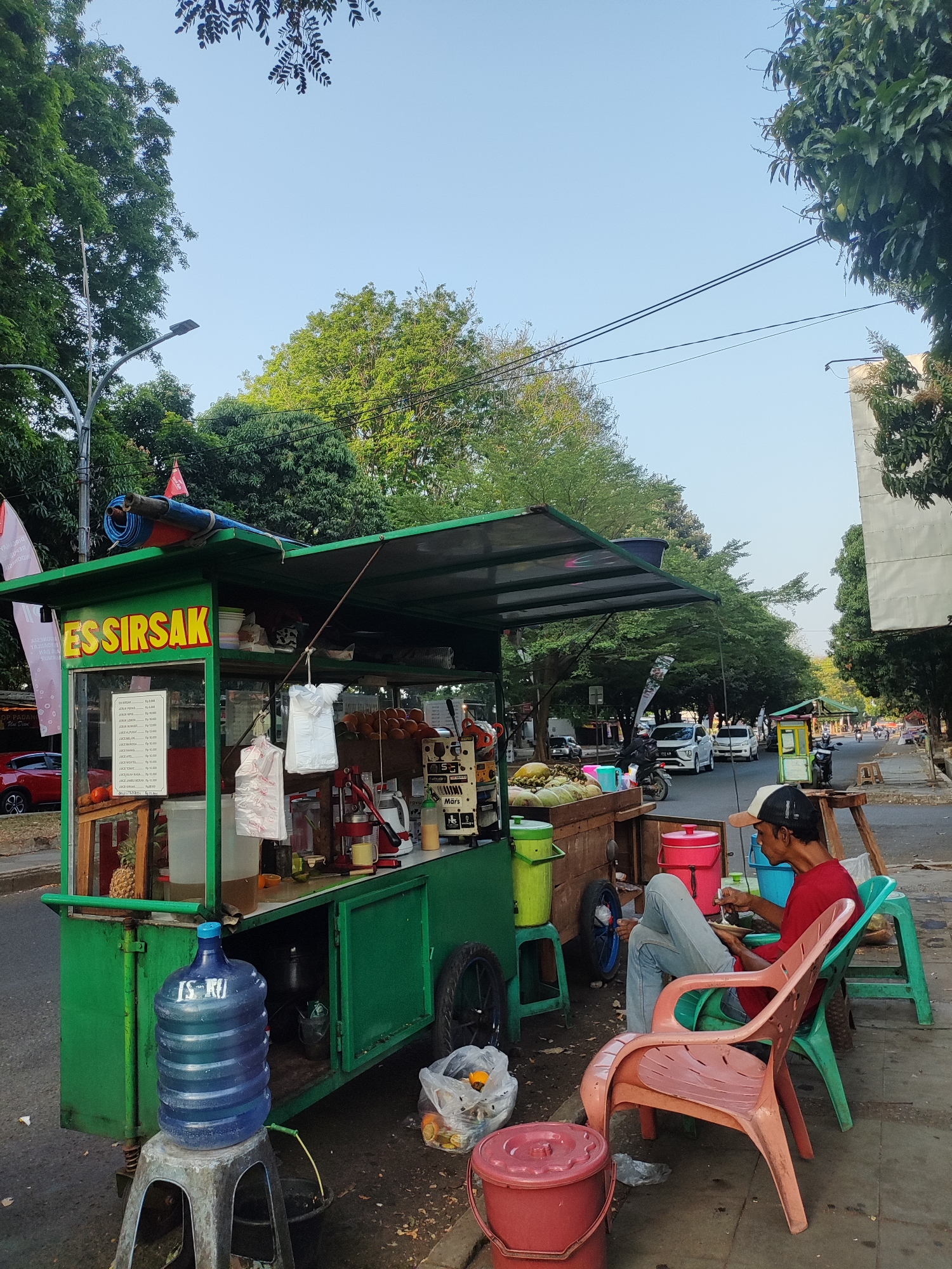 A man sits on a plastic chair next to a juice stand on a street in Cirebon, Indonesia. The juice stand, painted green, has a sign that reads "ES SIRSAK." The man, wearing a red shirt and blue jeans, appears to be eating or drinking from a small white dish. In front of him are several chairs, a large blue water dispenser, and a red bucket. The background shows a street with traffic, trees, and buildings. The sky is clear and blue.