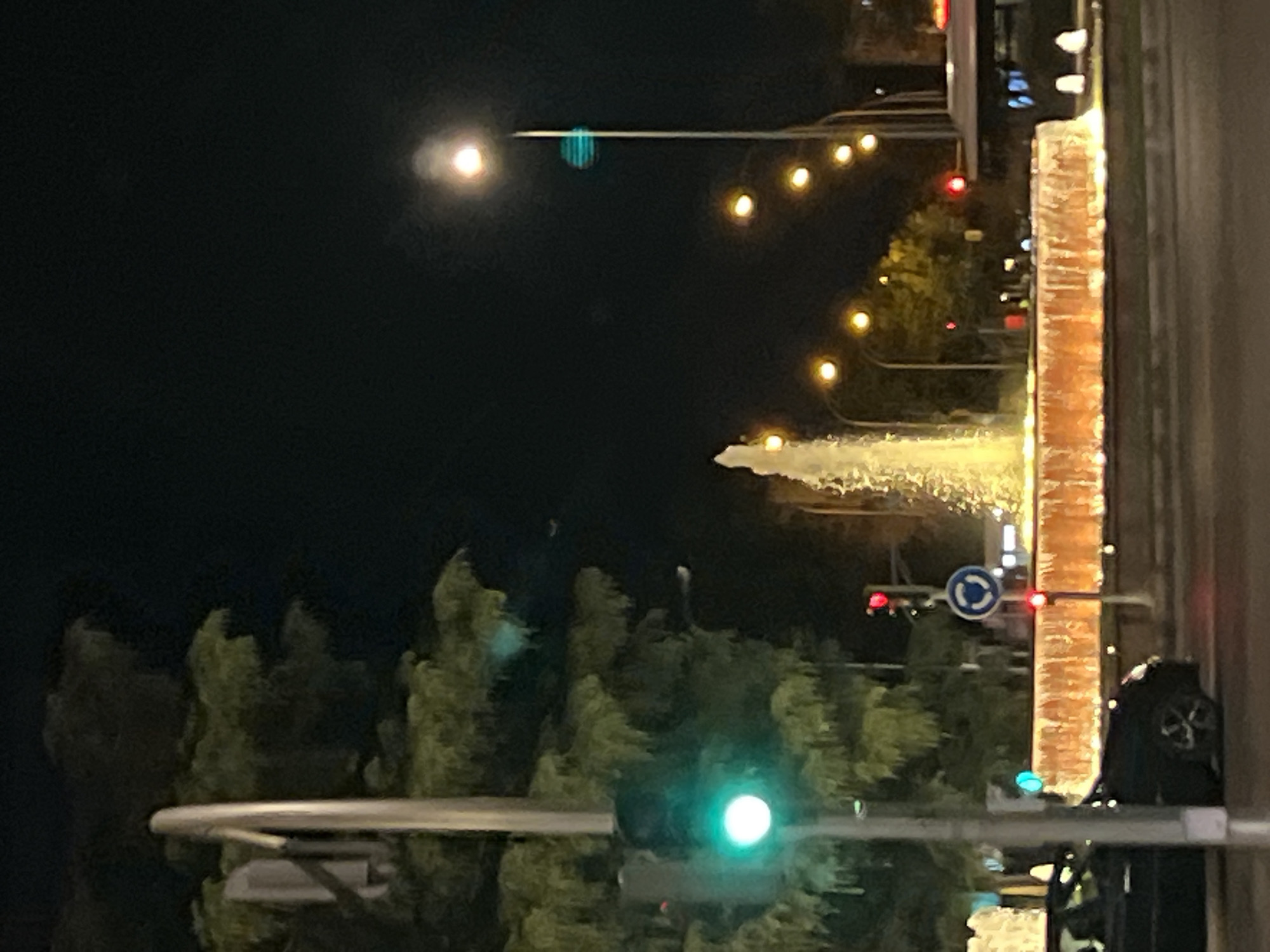The image depicts a night scene at a fountain in Madrid, Spain. In the foreground, a dark-colored car is partially visible on the left, driving past. A traffic light shows a green signal. Behind the car and traffic light, a large tree with lush green foliage is visible.
The central focus of the image is a prominent fountain with a strong jet of water illuminated by warm, yellow light. The fountain's basin is also brightly lit, appearing as a glowing, circular structure.
In the background, several streetlights cast a warm glow against the dark night sky. One tall streetlight on the right has a bright light at its top and a bluish-green light lower down. A circular road sign indicating a roundabout is visible near a red traffic light. Further in the background, the outline of a bus and some buildings can be seen. The overall impression is a well-lit urban setting at night, possibly a plaza or intersection with a decorative fountain.