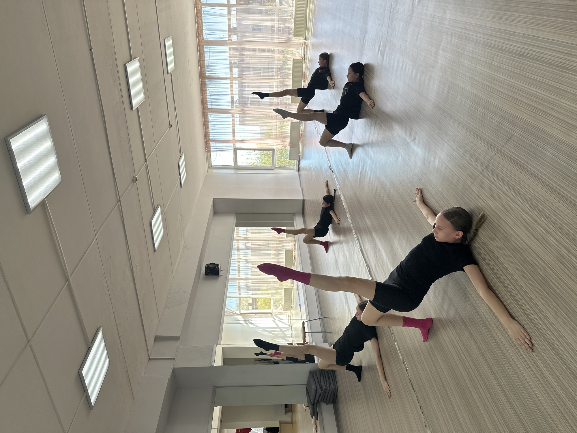 The image shows a dance studio with four young dancers practicing a routine on a light-colored, wood-patterned floor. They all wear black dance attire. One girl has bright pink socks, one has black socks, and the remaining dancers do not have socks. They are all lying on the floor, legs raised. The studio has a wall of windows letting in natural light and overhead fluorescent lighting. The location is Votkinsk, Russia.