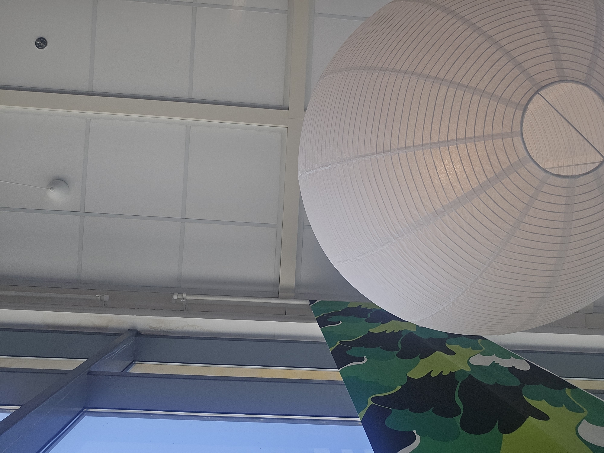 An indoor, close-up, low-angle shot captures a large, white, paper lantern hanging from a white ceiling. The lantern is spherical and ribbed with thin, gray lines radiating from the top. To the right of the lantern, a portion of a window with a blue sky is visible. Adjacent to the window, there is a section of wall with a green and black abstract pattern. The ceiling is made of white, square panels with thin gray grid lines. A thin, white electrical conduit runs vertically along the right side of the frame. The scene appears to be a well-lit interior space, possibly an office or a modern building. There are no people or animals present in the image. The specific location in Baronissi, Italy cannot be determined from the visual information provided.