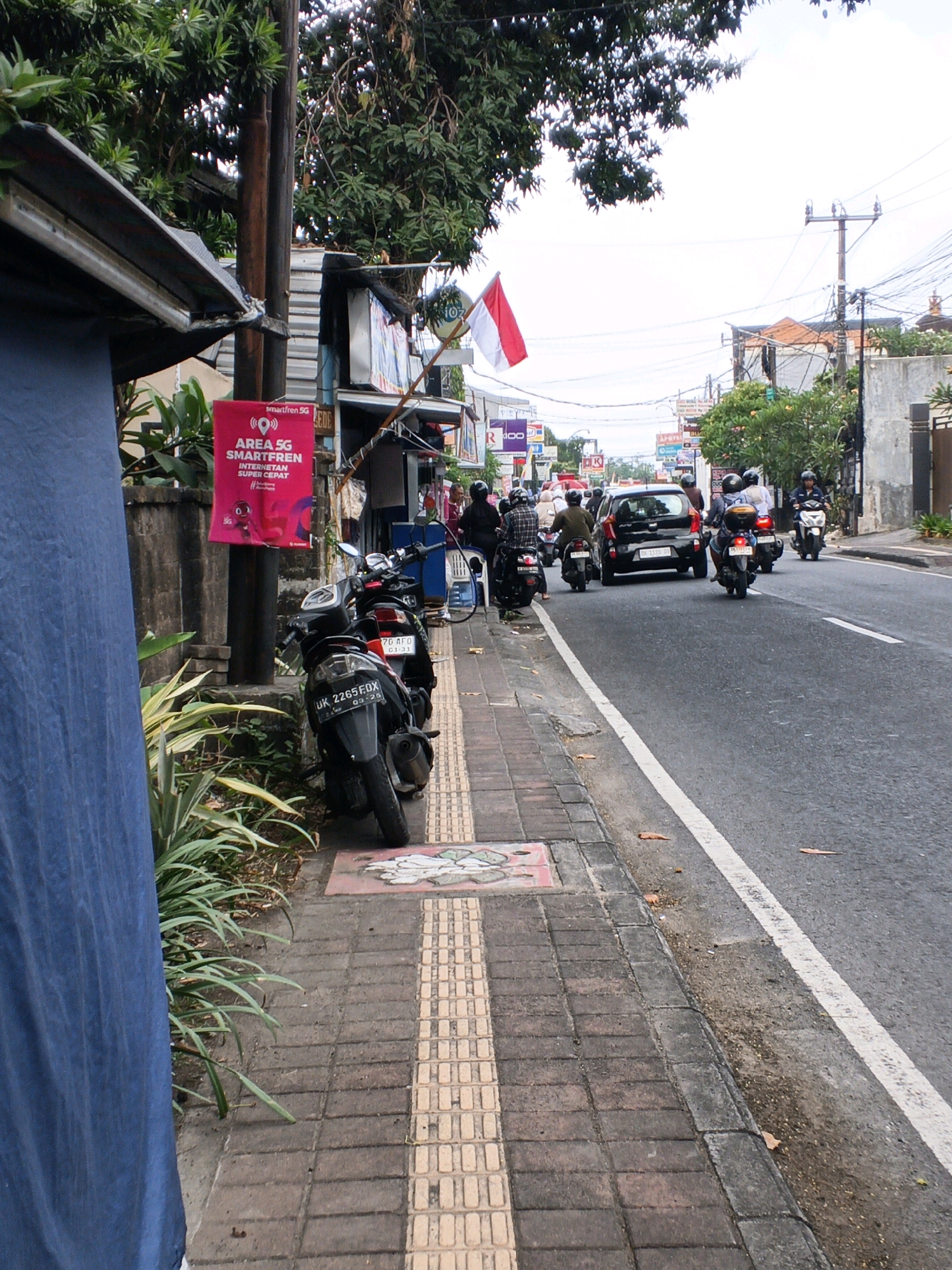 The image depicts a street scene in Denpasar, Indonesia, with a focus on a sidewalk and the adjacent road. Several motorcycles are parked on the sidewalk, and some are in motion on the road. A black car is also visible on the road. On the left side of the image, a blue tarpaulin covers part of a structure, and a pink sign advertising "AREA 5G SMARTFREN INTERNET SUPER CEPAT" is attached to a pole. A small plant with long, green leaves grows near the sidewalk. The road is paved and has a white line marking. In the background, buildings, power lines, and trees are visible. The sky is overcast. The Indonesian flag, red and white, is seen flying on a pole in the distance. The scene suggests a typical urban street environment in Denpasar, with an emphasis on local transportation and commercial advertisements.