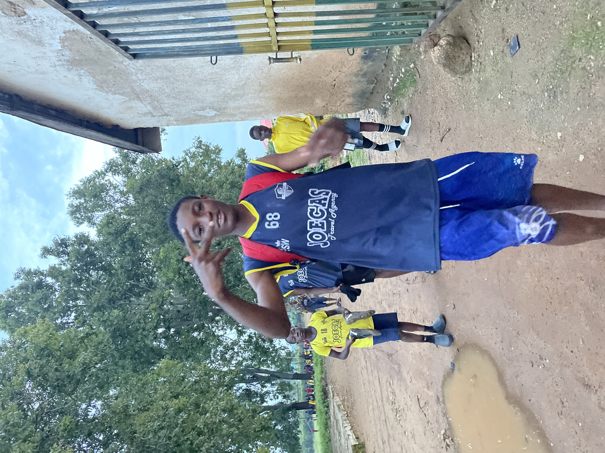 In Jos, Nigeria, on a dirt path, a group of young men, likely athletes, are walking. The foreground features a young Black male wearing a navy blue basketball jersey with yellow trim, number "68" visible on the front, and a red backpack. He is making a peace sign with his right hand. Behind him, another young Black male in a yellow t-shirt with "18" printed on it, holds a pair of dark-colored soccer cleats. Further back, a third young Black male in a bright yellow jacket and gray shorts walks past a textured, light-colored building with a metal gate. The environment is characterized by lush green trees, a cloudy sky, and a large puddle of muddy water on the path, suggesting recent rain. Other people and children are visible in the distance, engaged in activities on a grassy field. The scene depicts a casual, outdoor setting in what appears to be a community or school area.