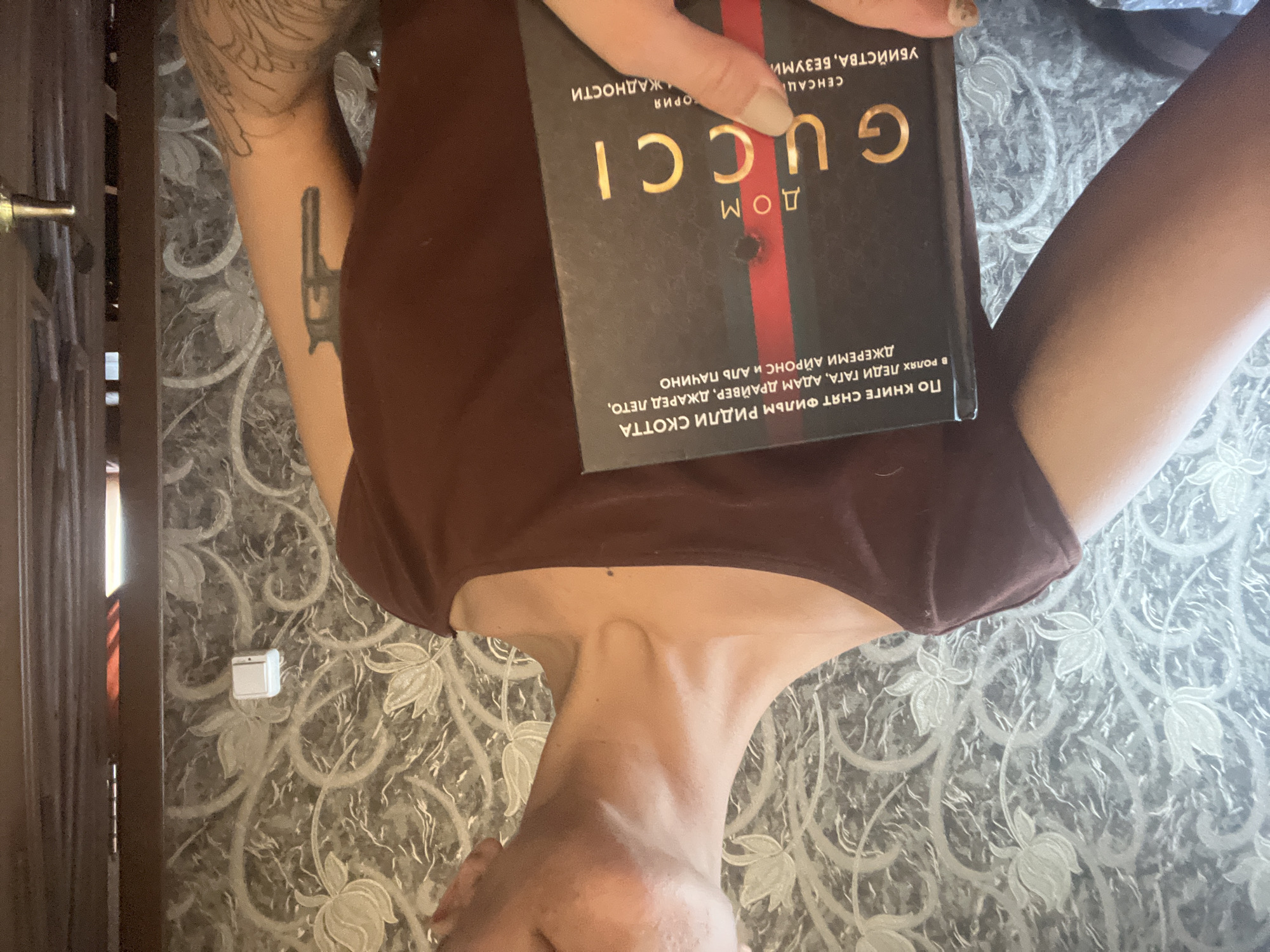 A person is holding a Russian edition of the book "House of Gucci," which is about the Gucci family. The person is lying on a surface with a floral pattern, possibly a carpet or bedspread. The person is wearing a brown top. The person has tattoos on their arm. A doorframe and a light switch can be seen in the background. The image was taken in Boguchar, Russia.