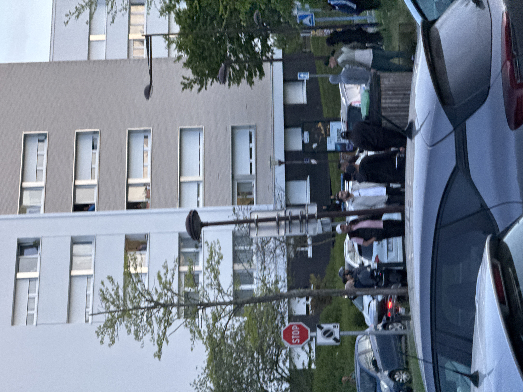 The image shows a street scene in Les Ulis, France. A modern, multi-story building with rectangular windows stands in the background. In the foreground, several parked cars are visible. A group of people are standing on the sidewalk between the cars and the building. A stop sign and streetlights are also present. The lighting suggests it's either early morning or late afternoon.