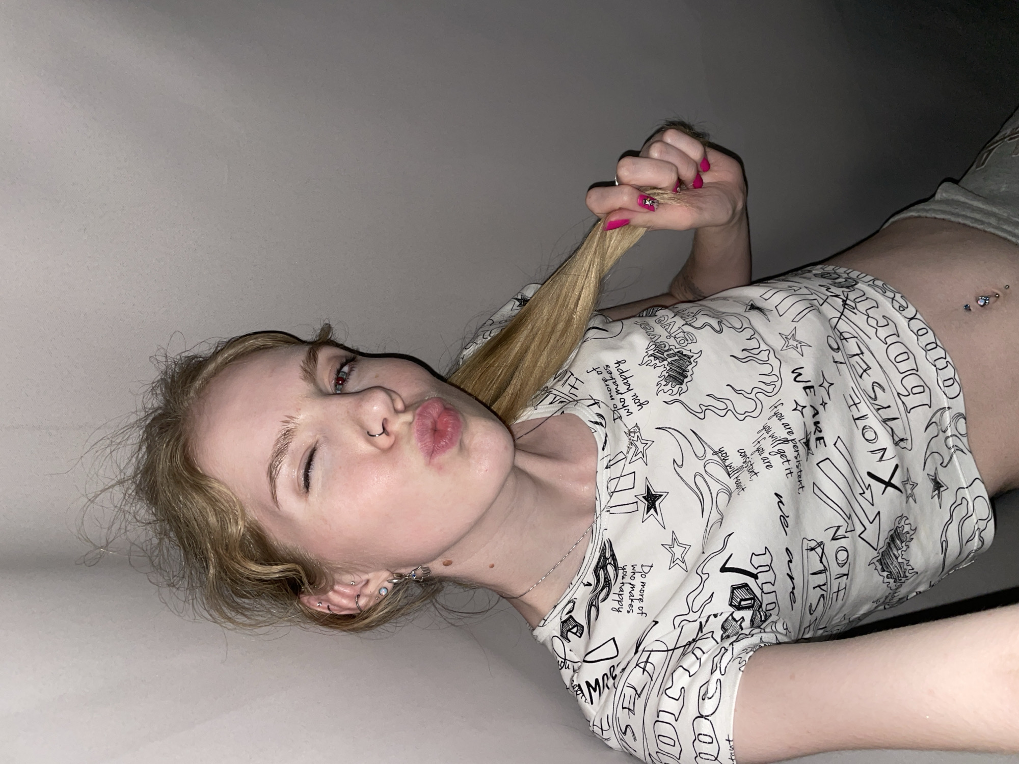 A fair-skinned woman with blonde hair and pink nail polish is pulling her hair in the upper right-hand corner of the image. She is wearing a white, printed crop top and gray shorts. She has several ear piercings on her left ear, a nose ring, and a belly button piercing. Her lips are puckered, and her left eye is squinted. The background is a plain, light gray color. There is no indication of the city of Izhevsk, Russia, or any specific location.