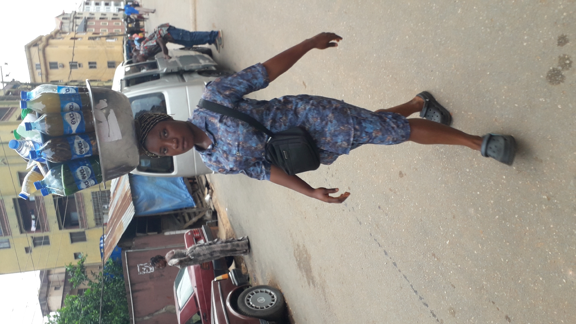 The image captures a woman walking on a street in Onitsha, Nigeria. She balances a large metal tray on her head, which is filled with bottles labeled "Eva." She is wearing a blue patterned shirt, shorts, and black Crocs, with a small black bag slung across her shoulder. The background includes buildings, vehicles (a maroon car and a silver van), and other people. The street is paved, and the overall atmosphere is that of a bustling urban scene.