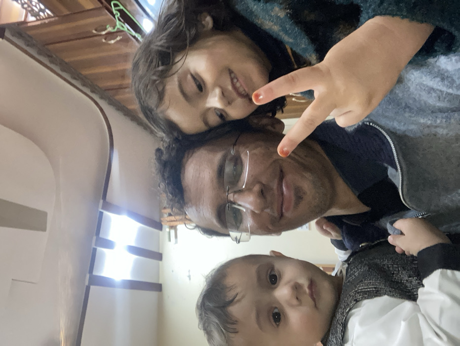 A rotated selfie features a man with glasses, a young girl, and a toddler. The girl, positioned above the man, smiles and holds up a peace sign. The man looks directly at the camera with a slight smile. The toddler is placed at the bottom of the frame. The background is a simple interior with a white wall and some visible trim. The overall composition suggests a casual, familial moment captured on camera.