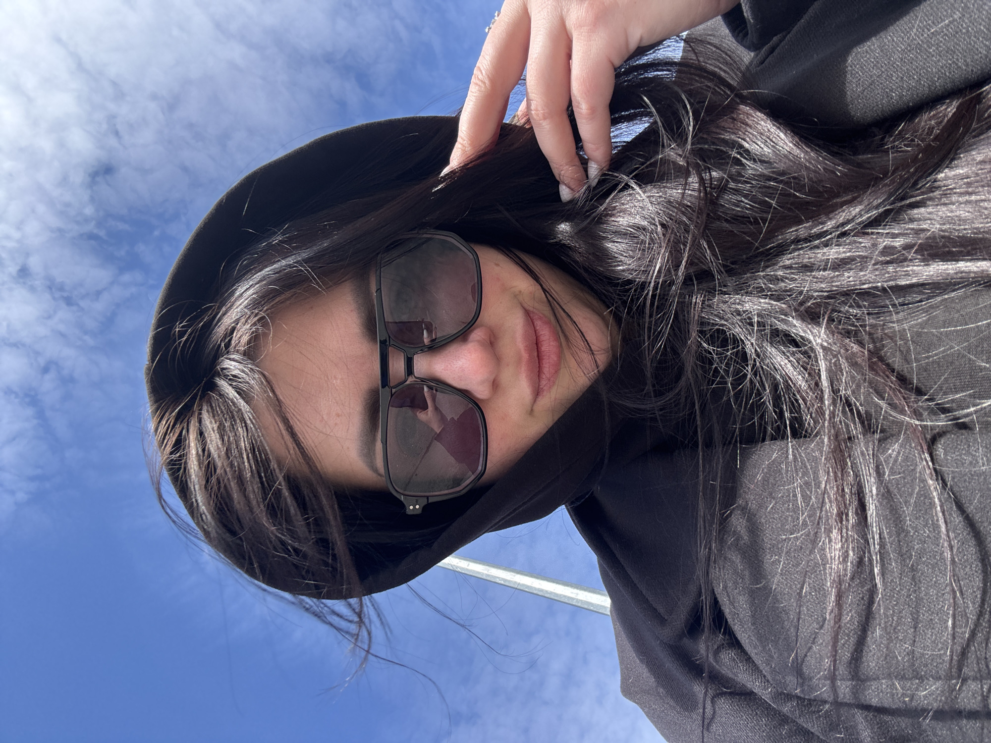 A young woman wearing dark sunglasses and a black hooded sweatshirt is pictured from a low angle against a blue sky with scattered clouds. She is touching her hair with her left hand, which has long white fingernails. A metal pole is visible in the lower left corner of the image. The exact location is unknown.
