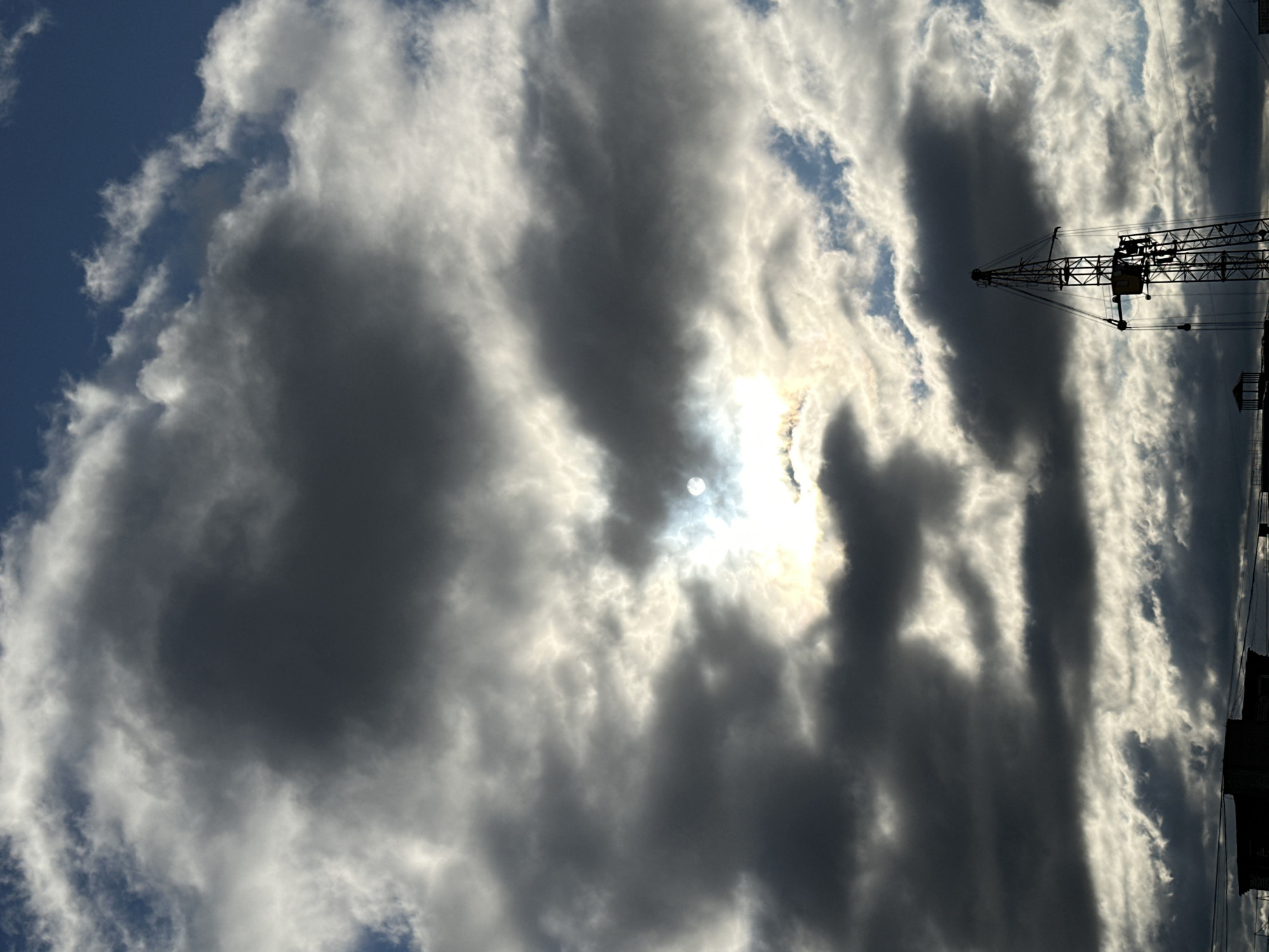 The image shows a cloudy sky with the sun peeking through. A construction crane is visible against the sky on the right side of the frame. The location is Chaikovsky, Russia.