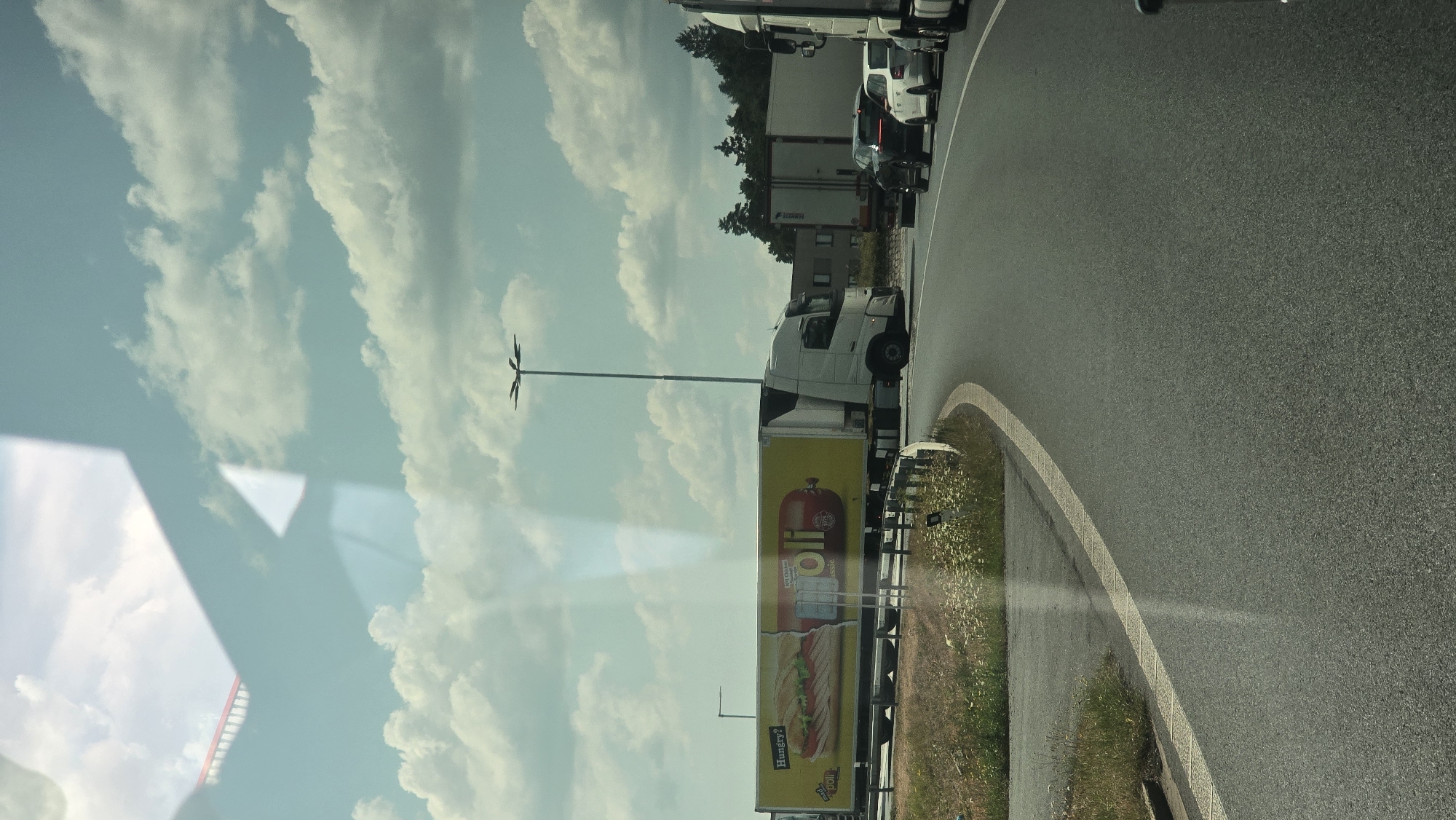 The image shows a highway intersection in Bor, Czechia, on a cloudy day. In the foreground, a gray asphalt road curves to the right. To the left of the road is a grassy embankment with white wildflowers. A large white trailer truck with a yellow advertisement featuring a sandwich and a sausage with the brand name "oli" is driving on the road. Behind it, other vehicles, including a white car and a black SUV, are also visible. In the background, a tall street lamp stands near a line of trees. The scene is captured through a reflective surface, likely a car window, as evidenced by a shadow and reflections of the sky and clouds.