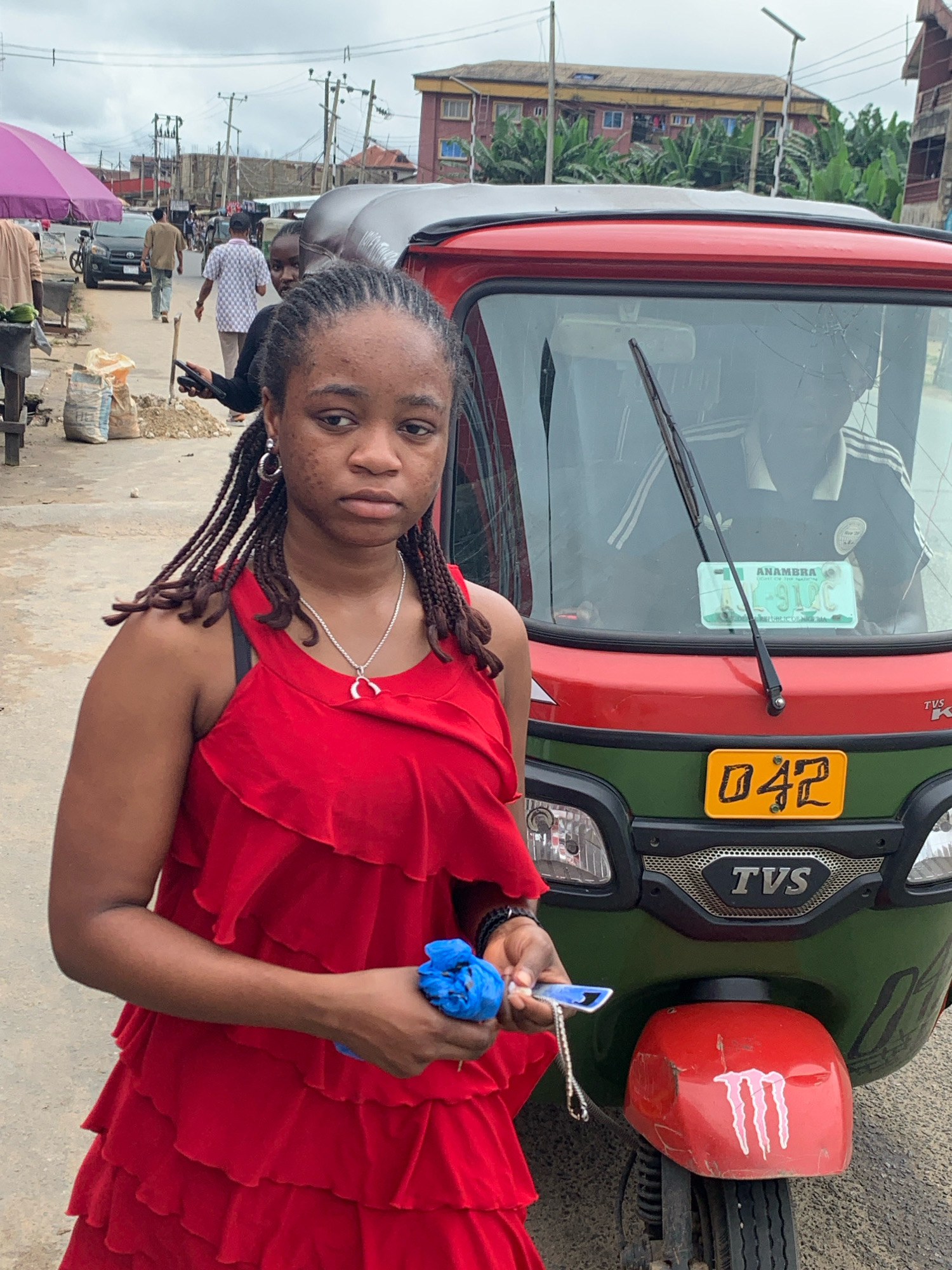 The image shows a young Black woman in the foreground, facing the camera with a neutral expression. She is wearing a red, layered, ruffled dress and a silver necklace with a heart-shaped pendant. Her hair is braided and falls over her shoulders. She is holding a blue object and a keychain in her right hand. To her right, a red and green auto-rickshaw is parked. The auto-rickshaw has a cracked windshield, a license plate that reads "ANAMBRA L-91 BC", and a yellow license plate on the front that reads "042". The backdrop depicts a street scene in Umuode, Nigeria, with buildings, power lines, and other people in the distance. A pink umbrella and a black car are visible to the left.