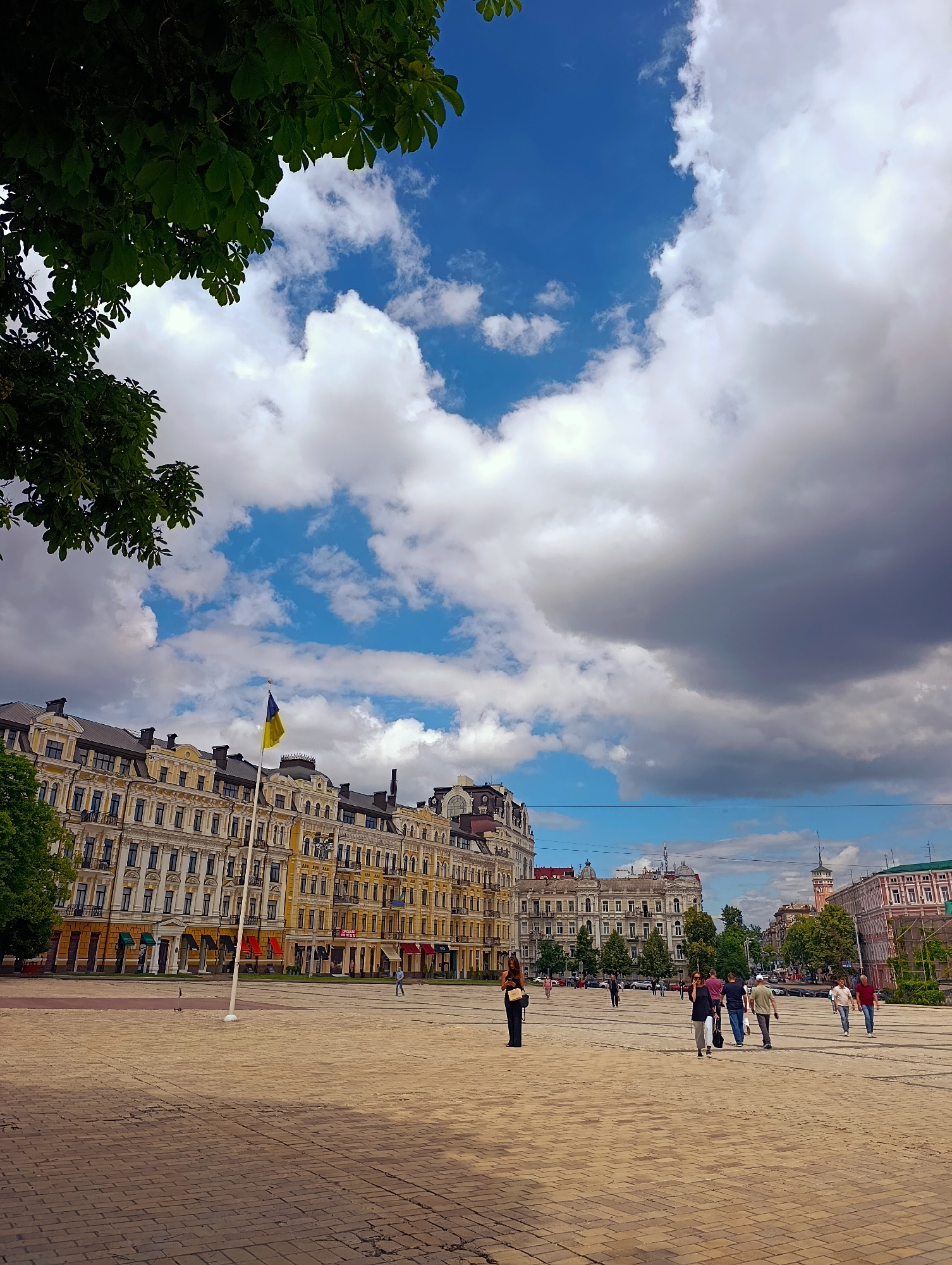 The media file depicts an outdoor scene in a European-style city square, likely Kyiv, Ukraine, given the presence of the Ukrainian flag flying from a flagpole. The sky is a mix of bright blue with scattered white clouds, and some darker, more ominous clouds suggest approaching weather. Lush green leaves from a tree frame the top left of the image, casting a shadow over part of the scene.
The foreground is a wide, paved plaza, made of light-colored bricks laid in a pattern. Several people are scattered across the plaza, walking in different directions. Their attire suggests a casual daytime activity, possibly sightseeing or moving between locations. The architecture of the buildings lining the square is ornate and historic, featuring yellow and beige facades with many windows and decorative elements. A Ukrainian flag, predominantly blue and yellow, is prominently displayed on a tall flagpole in front of the buildings.
The time of day appears to be daytime, given the brightness and the shadows. The weather seems to be variable, with patches of sunshine interspersed with heavier cloud cover. The overall mood is one of everyday life in a public space, with people going about their business against a backdrop of impressive historical architecture. There is no visible text in the image.
