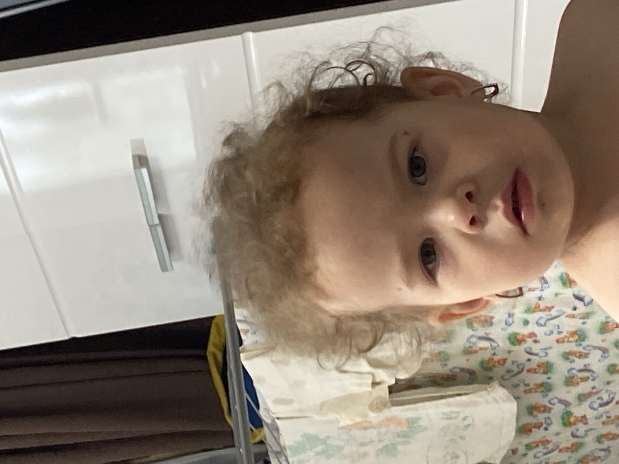 A young child with curly, light brown hair is positioned in the foreground of the image. The child's face is turned slightly to the right, with their gaze directed towards the camera. They have dark eyes and visible ear piercings. In the background, a white dresser with a horizontal handle is visible, along with dark brown curtains to the left and a rack of patterned fabric items. The overall setting appears to be indoors, likely a bedroom or living area.